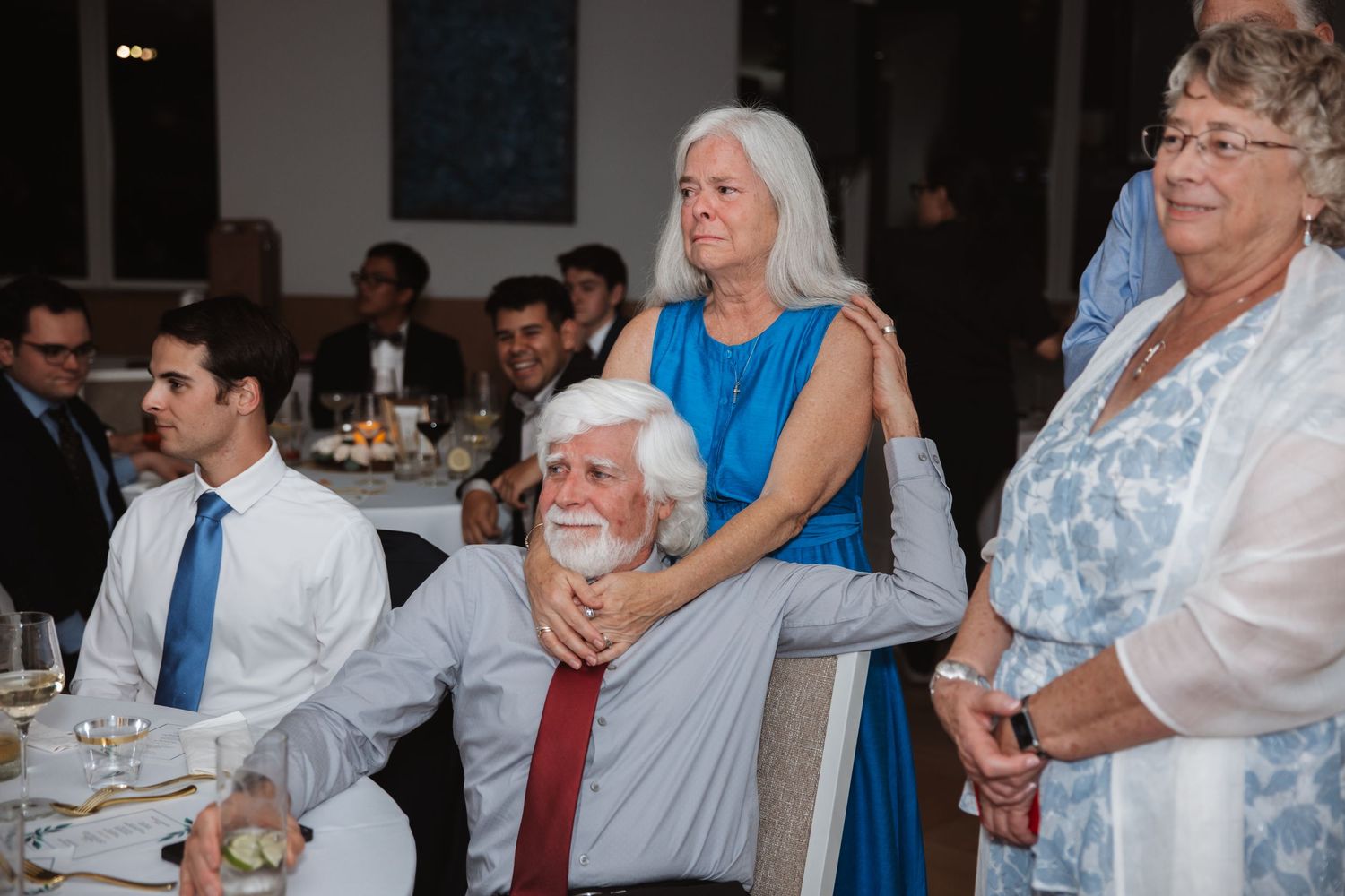 Wedding guests watching ceremony, including elderly couple embracing in blue and white attire.