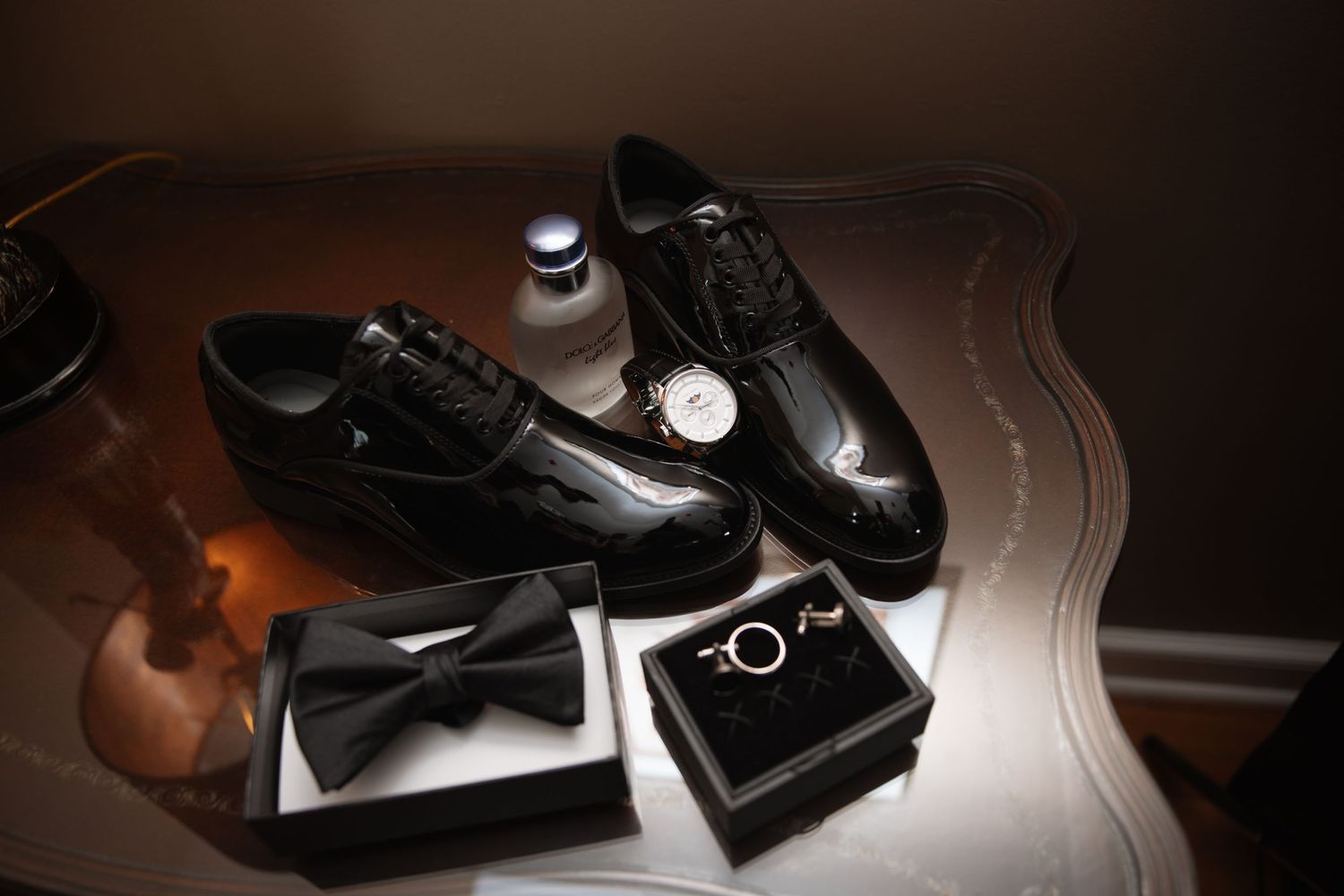Groom's patent leather shoes, bow tie, watch, and wedding rings arranged on table.
