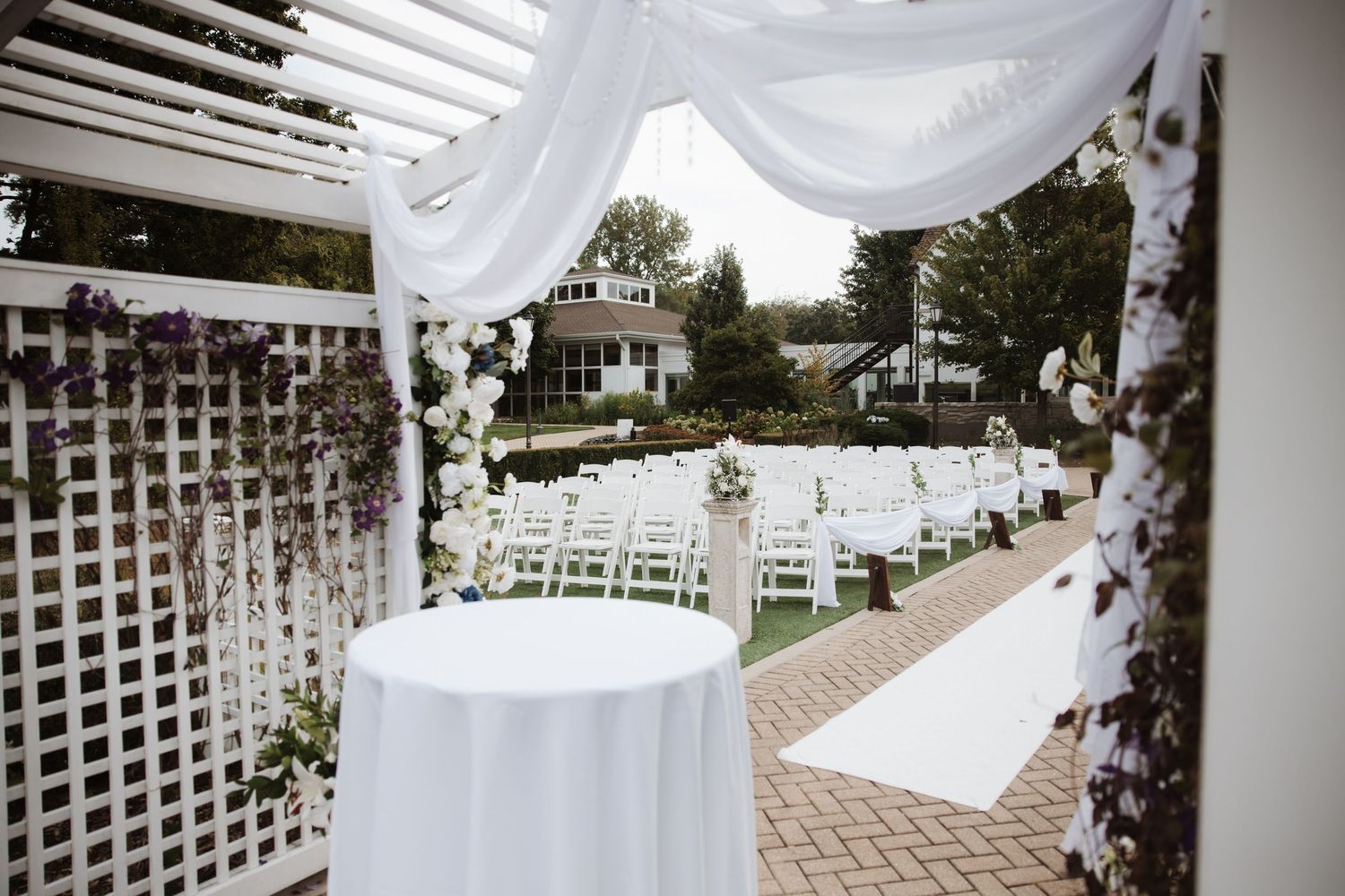 White lattice wedding arbor with floral arrangements and white draped fabric overlooking outdoor ceremony chairs.