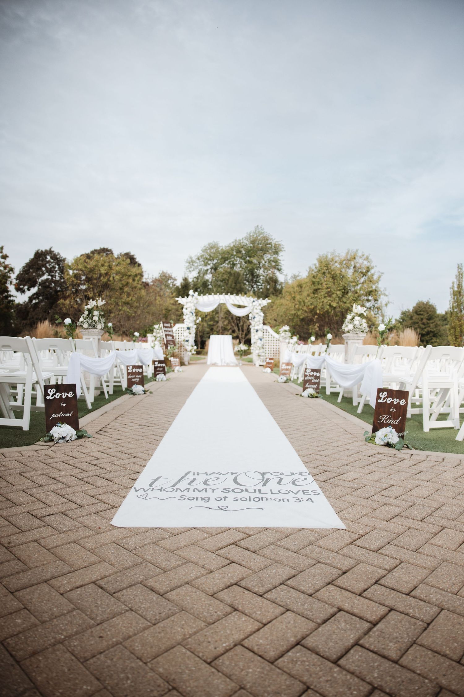 Wedding aisle runner with Song of Solomon scripture, white chairs, and floral arch in outdoor venue.