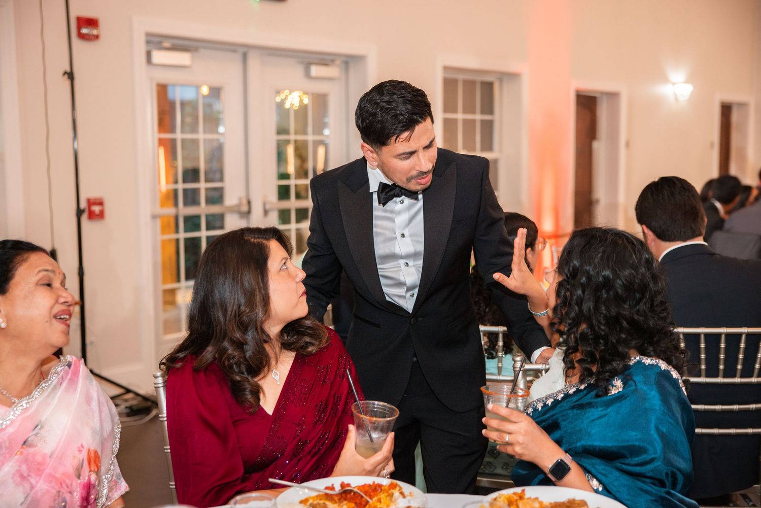 Man in tuxedo greeting women at elegant wedding reception dinner table.