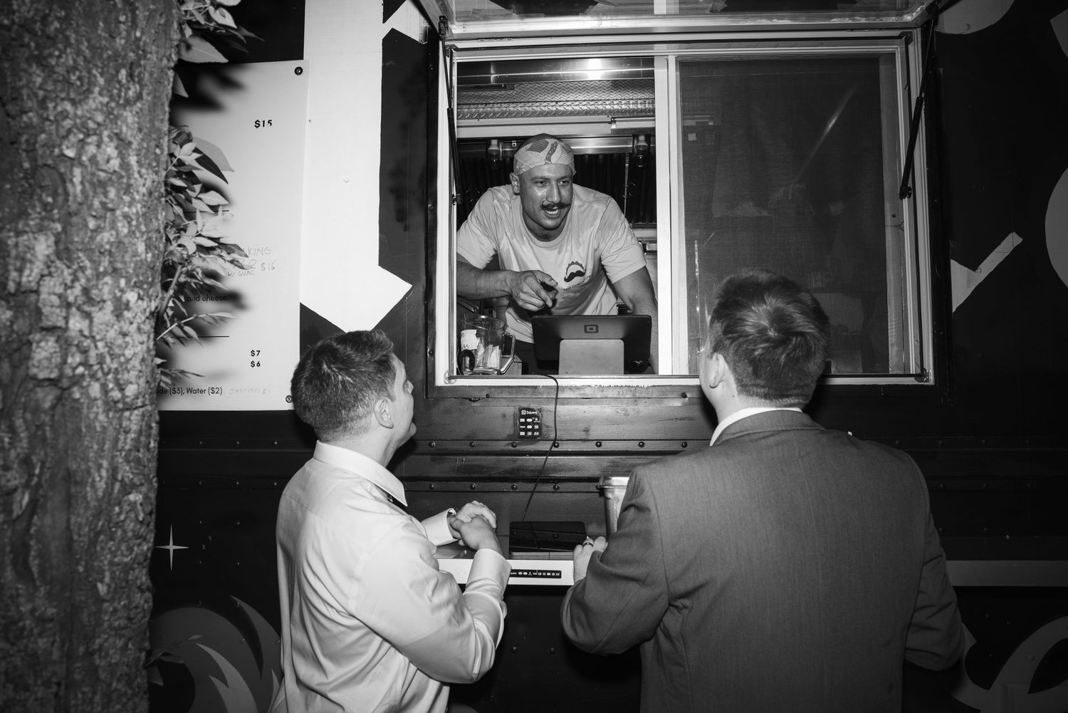 Black and white photo of two men ordering from a food truck window at night.