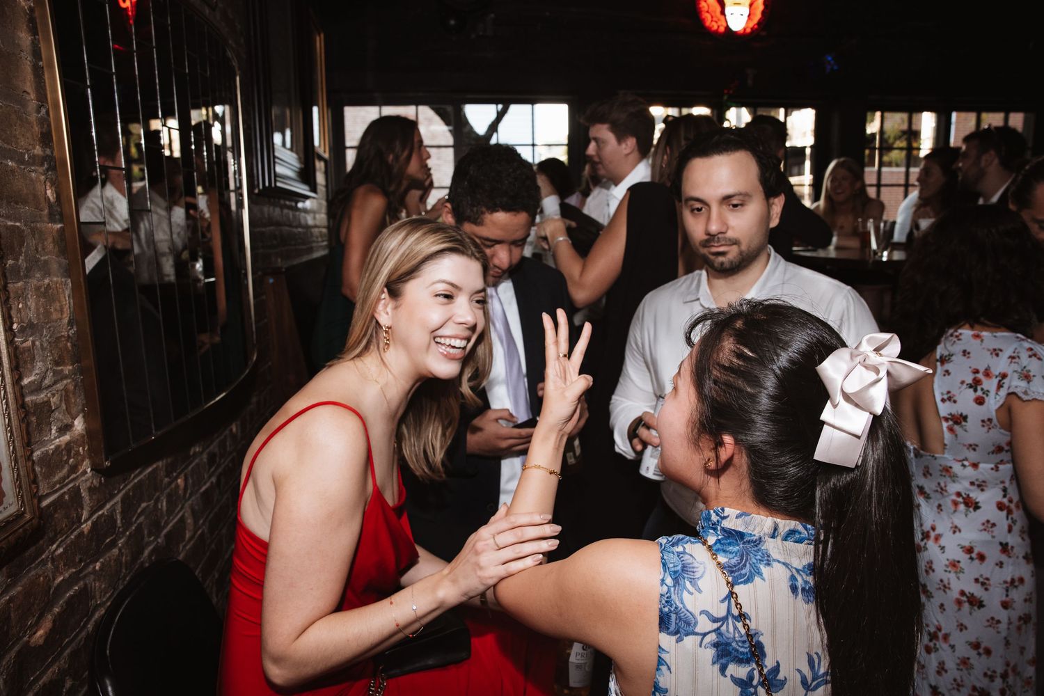 Two women in formal attire laugh and interact at a lively indoor wedding reception party.