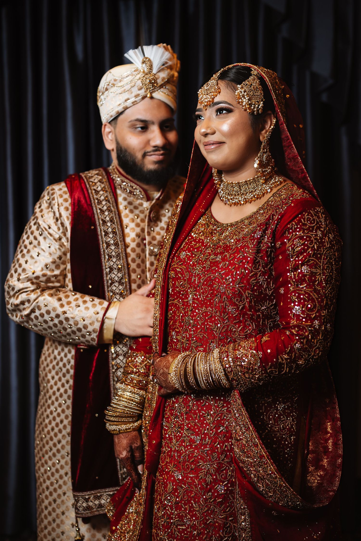 South Asian bride and groom in traditional red and gold wedding attire posing elegantly against dark backdrop.