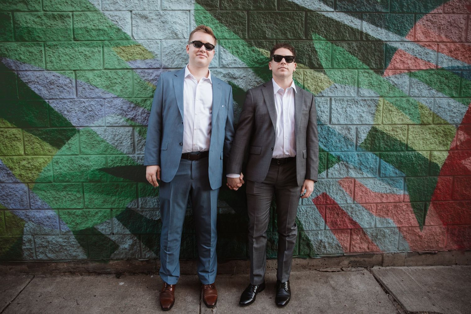 Two men in suits holding hands in front of a colorful geometric mural wall.