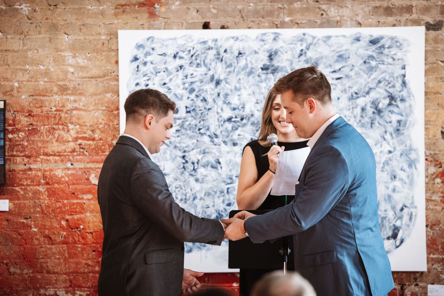Two grooms exchange rings with a female officiant during their wedding ceremony in an art gallery.