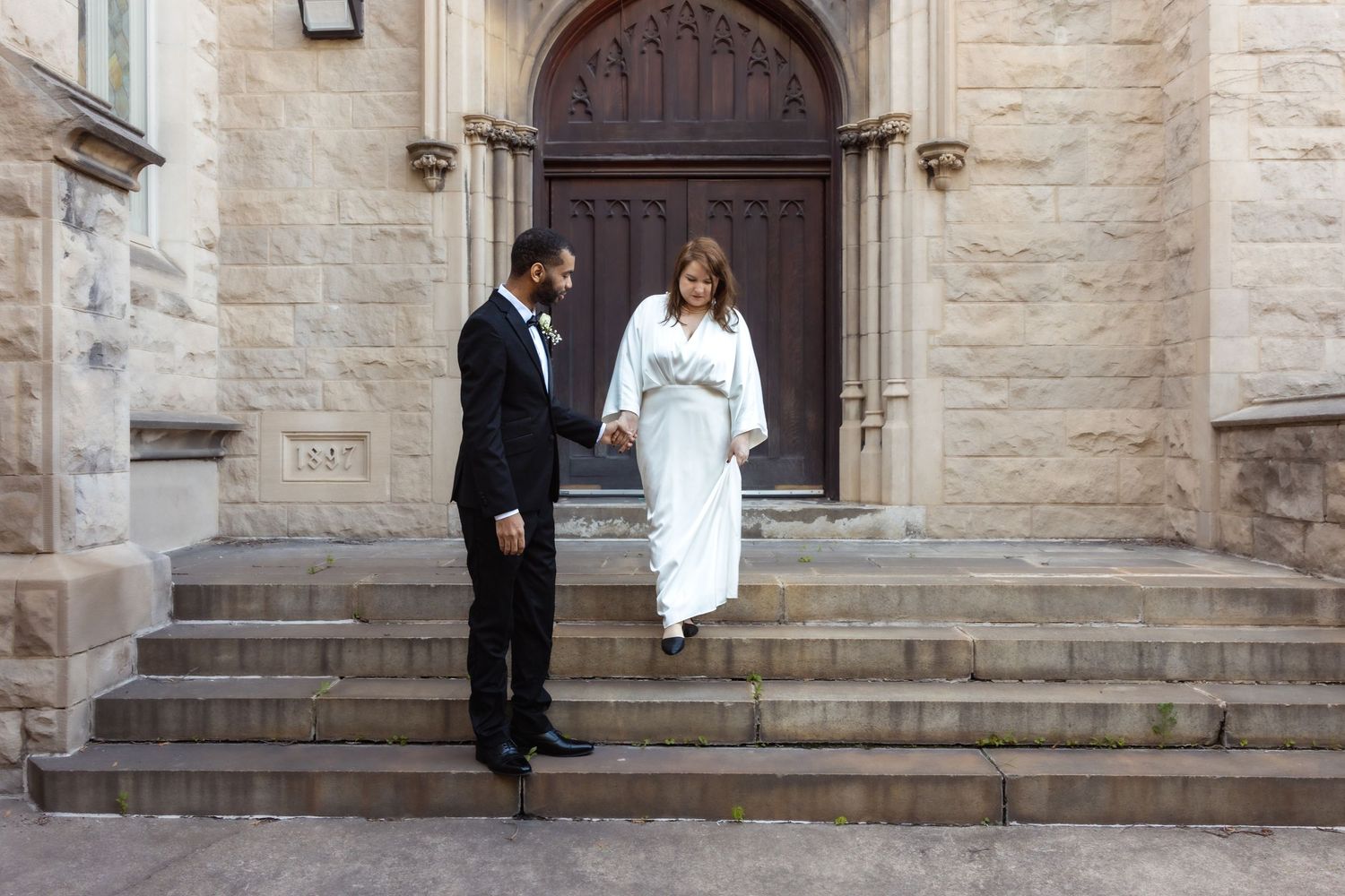 Bride and groom holding hands walking down stone church steps in wedding attire.