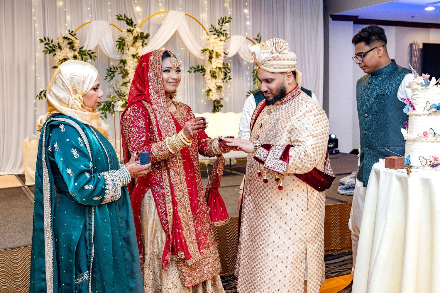 A bride in red lehenga and groom in cream sherwani exchange rings during a traditional South Asian wedding ceremony.