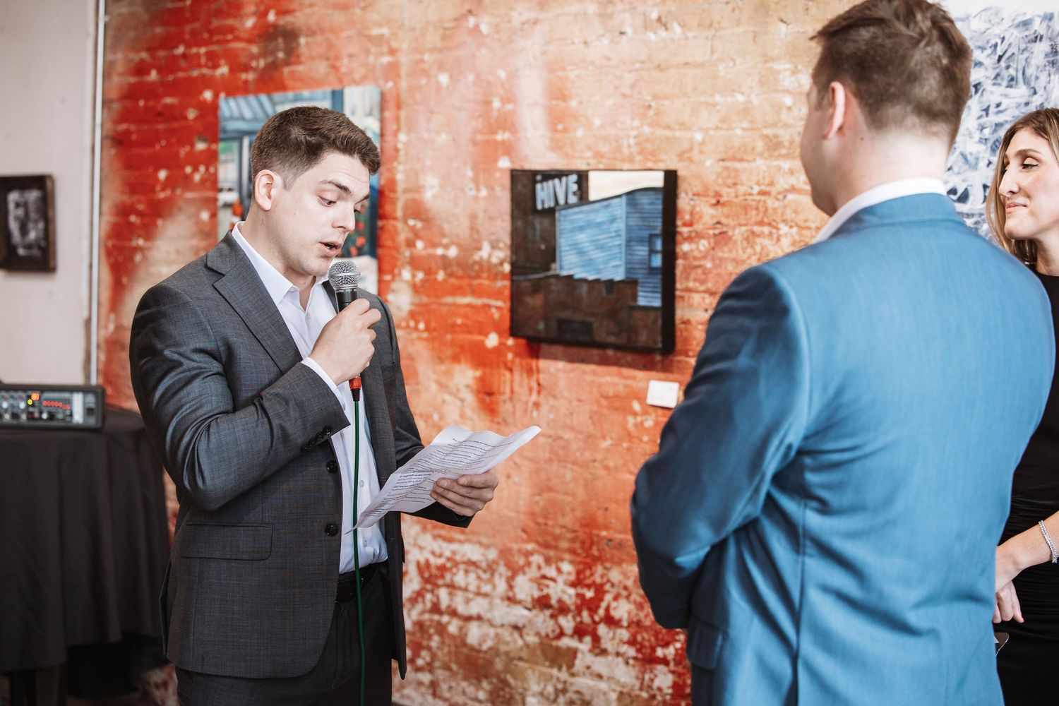 Man in a dark suit speaking into a microphone while holding papers at an indoor event near a brick wall.