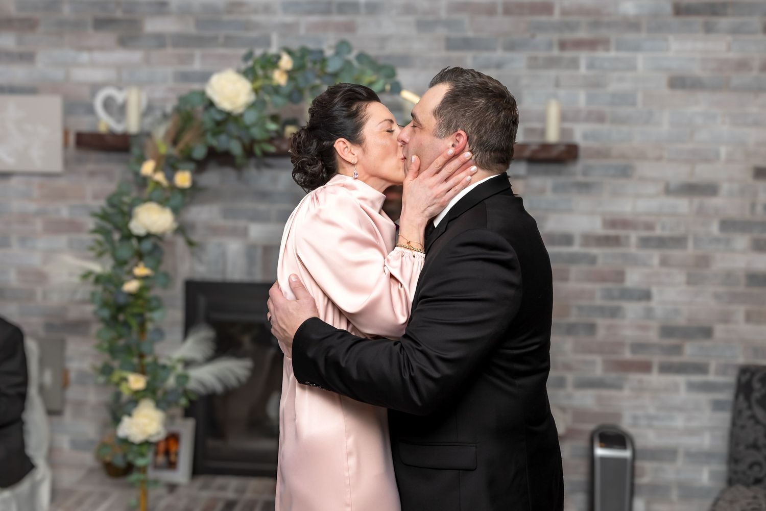 Newlywed couple shares first kiss in front of brick fireplace decorated with floral wedding arch.