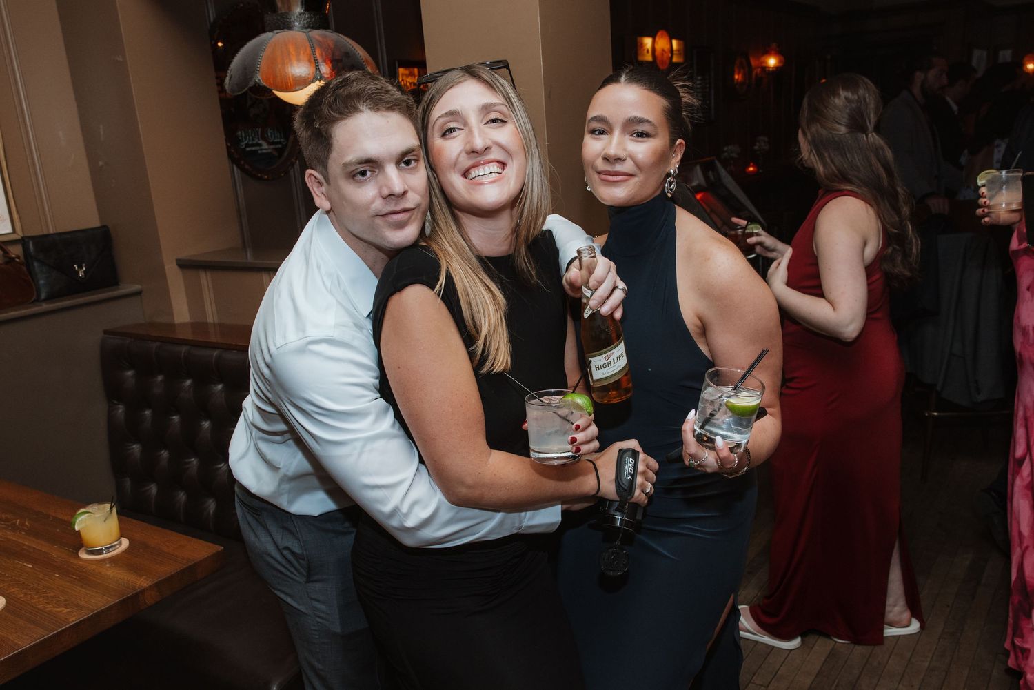 Three young wedding guests pose together holding cocktails at an indoor reception venue.