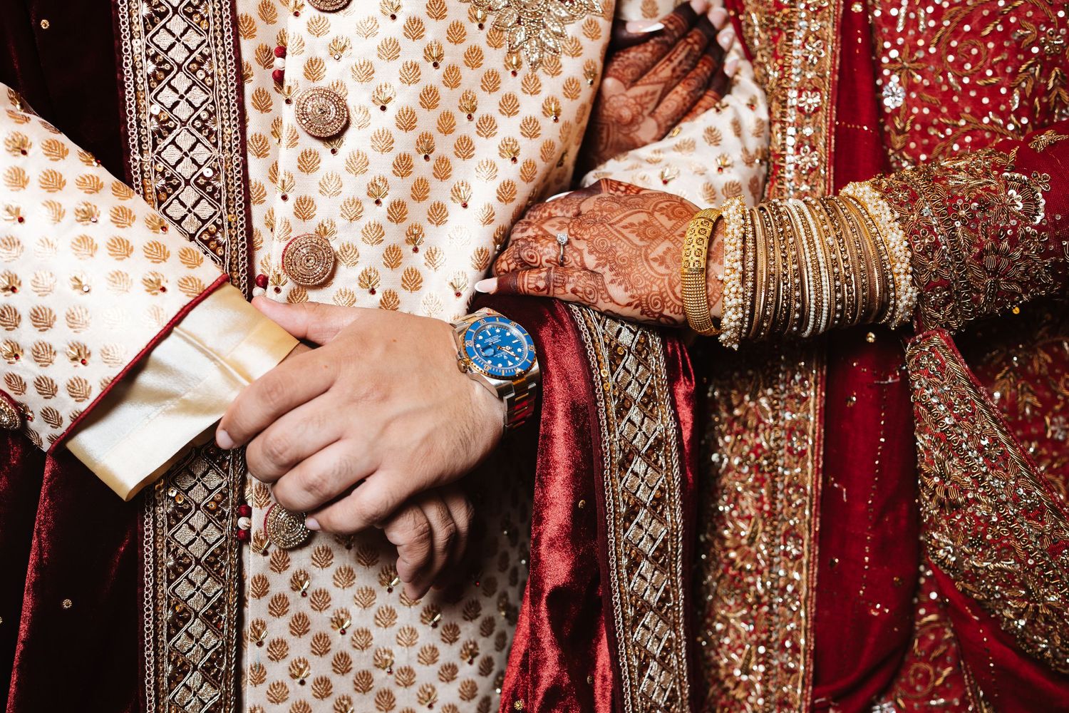 Close-up of Indian wedding couple in traditional attire, bride with mehndi and bangles, groom wearing blue watch.