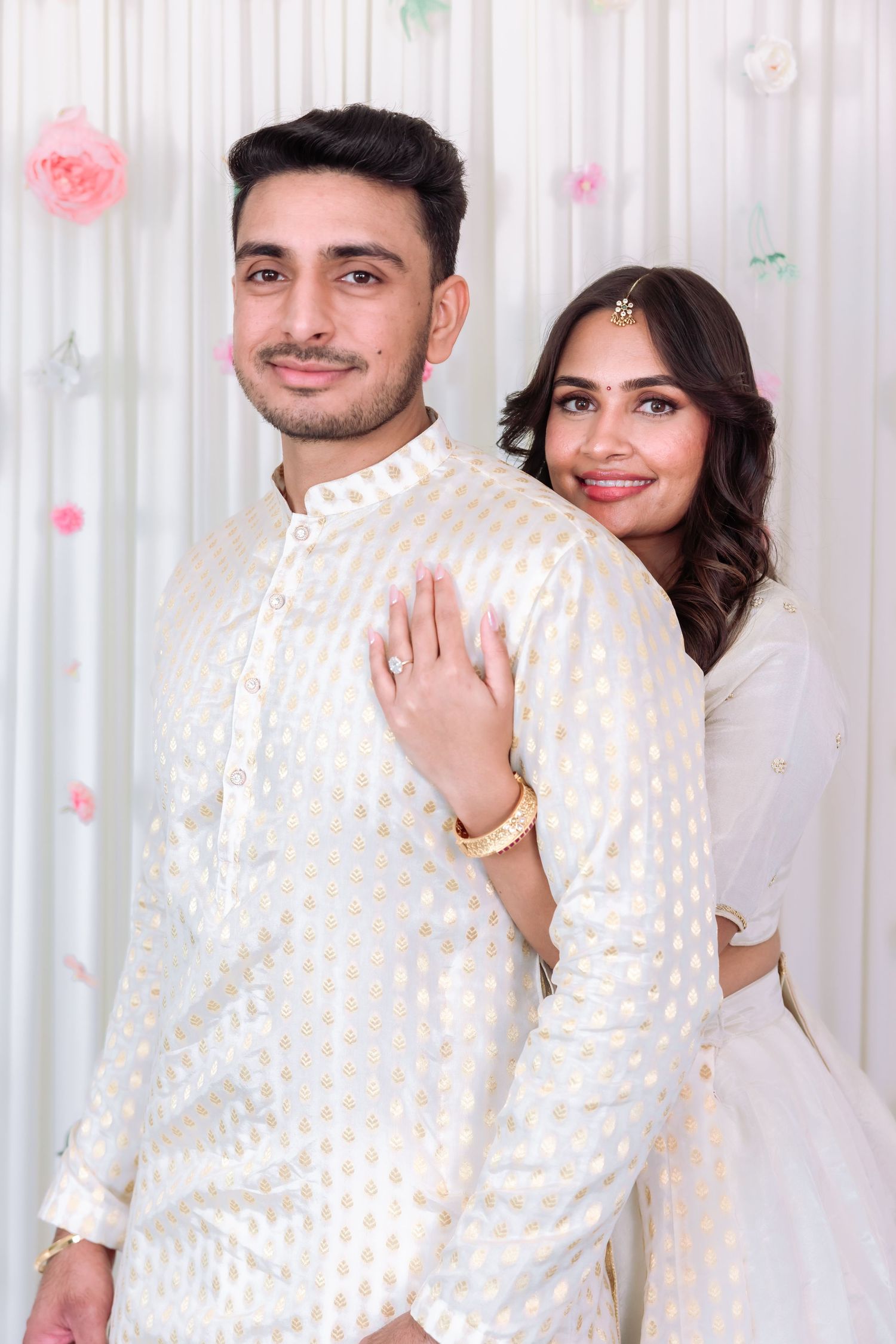 Indian couple in white traditional attire pose together at a floral decorated celebration backdrop.