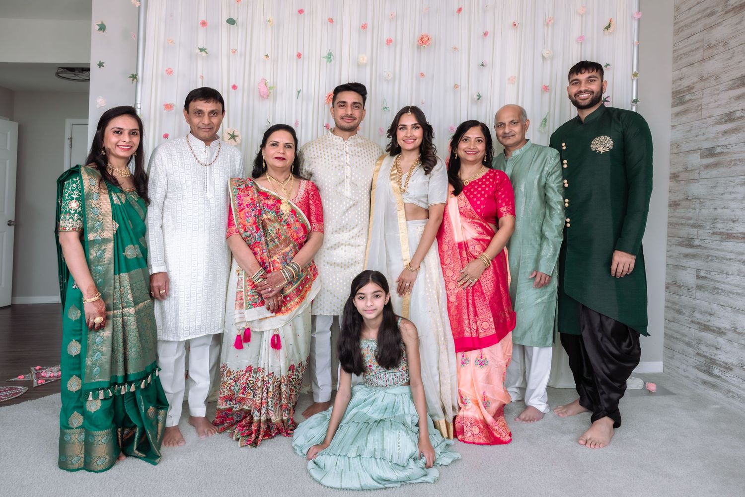 Indian wedding family group photo with members dressed in colorful traditional attire before floral backdrop.