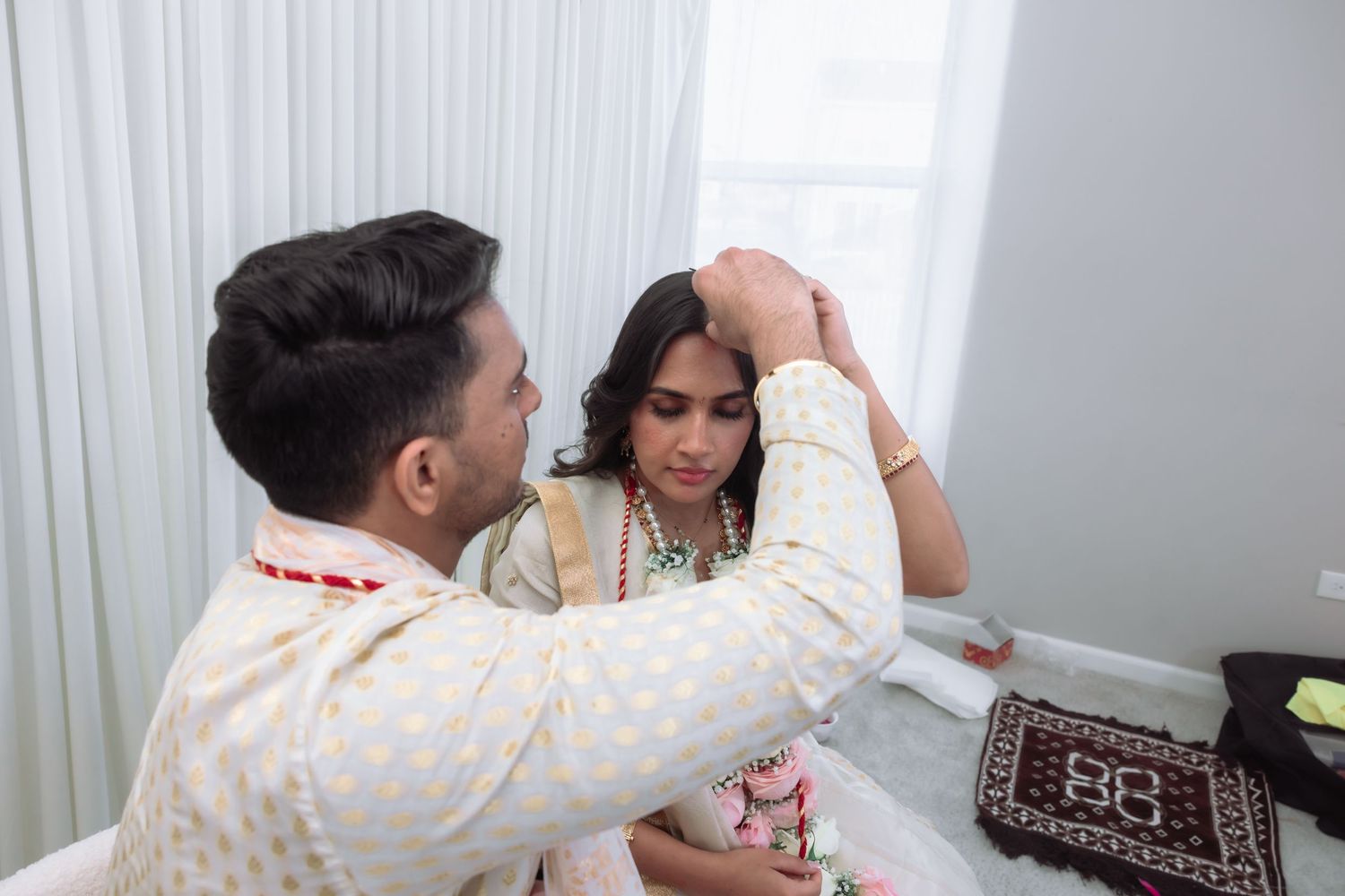 Groom adorns bride with maang tikka jewelry during intimate Hindu wedding ceremony indoors.