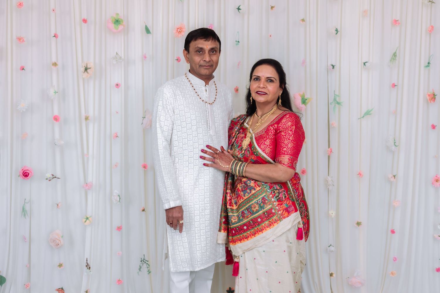 Indian couple in traditional festive attire smiling together against a white floral decorated backdrop.