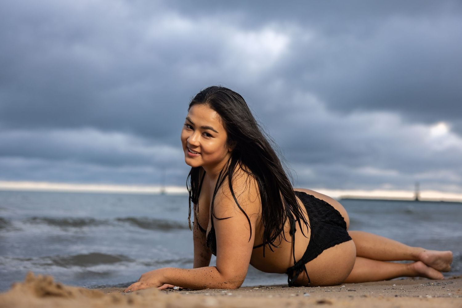 Smiling woman in black bikini posing on wet sand at beach with dramatic cloudy sky and ocean behind her.