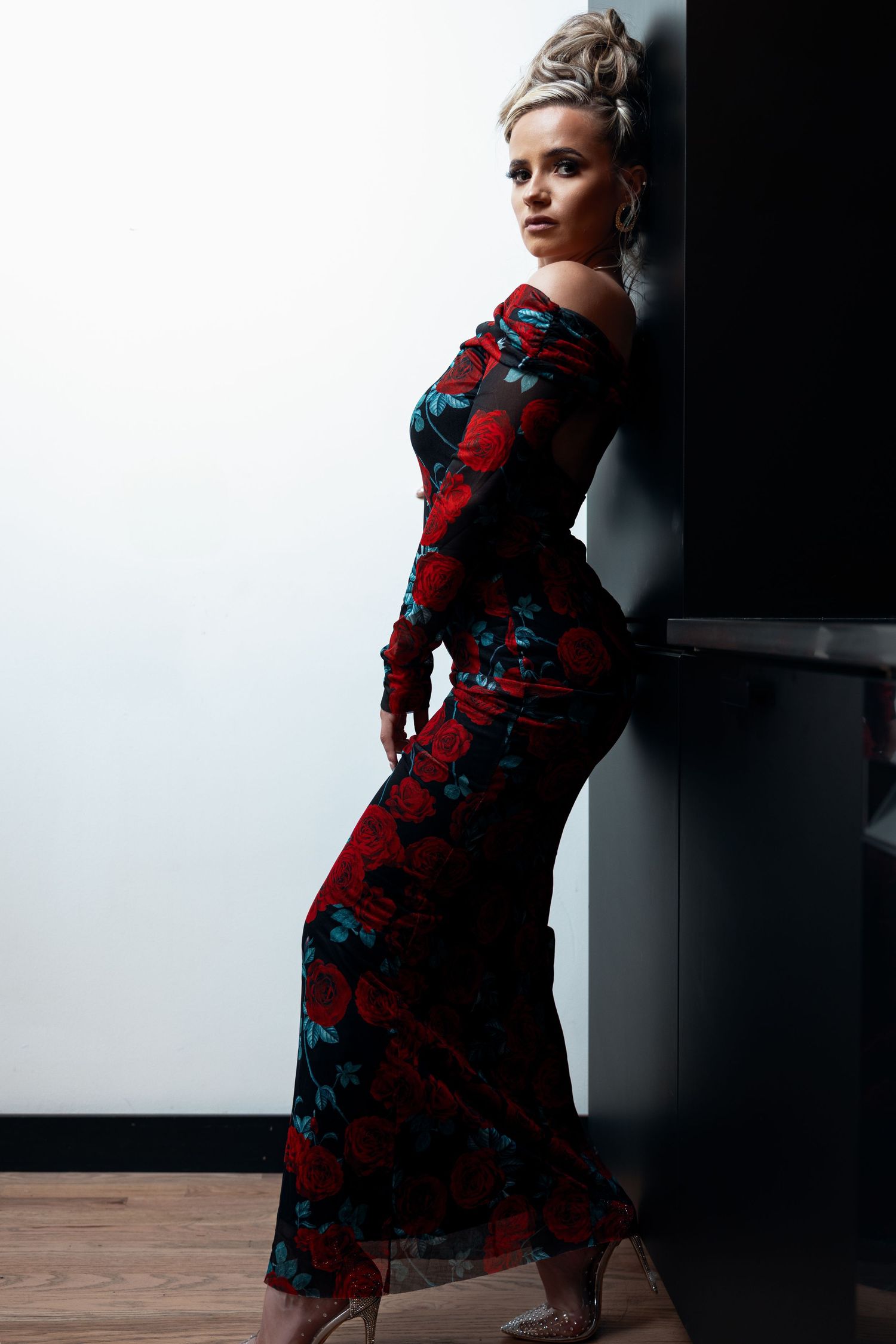 Woman in red rose print off-shoulder maxi dress posing sideways against dark wall.