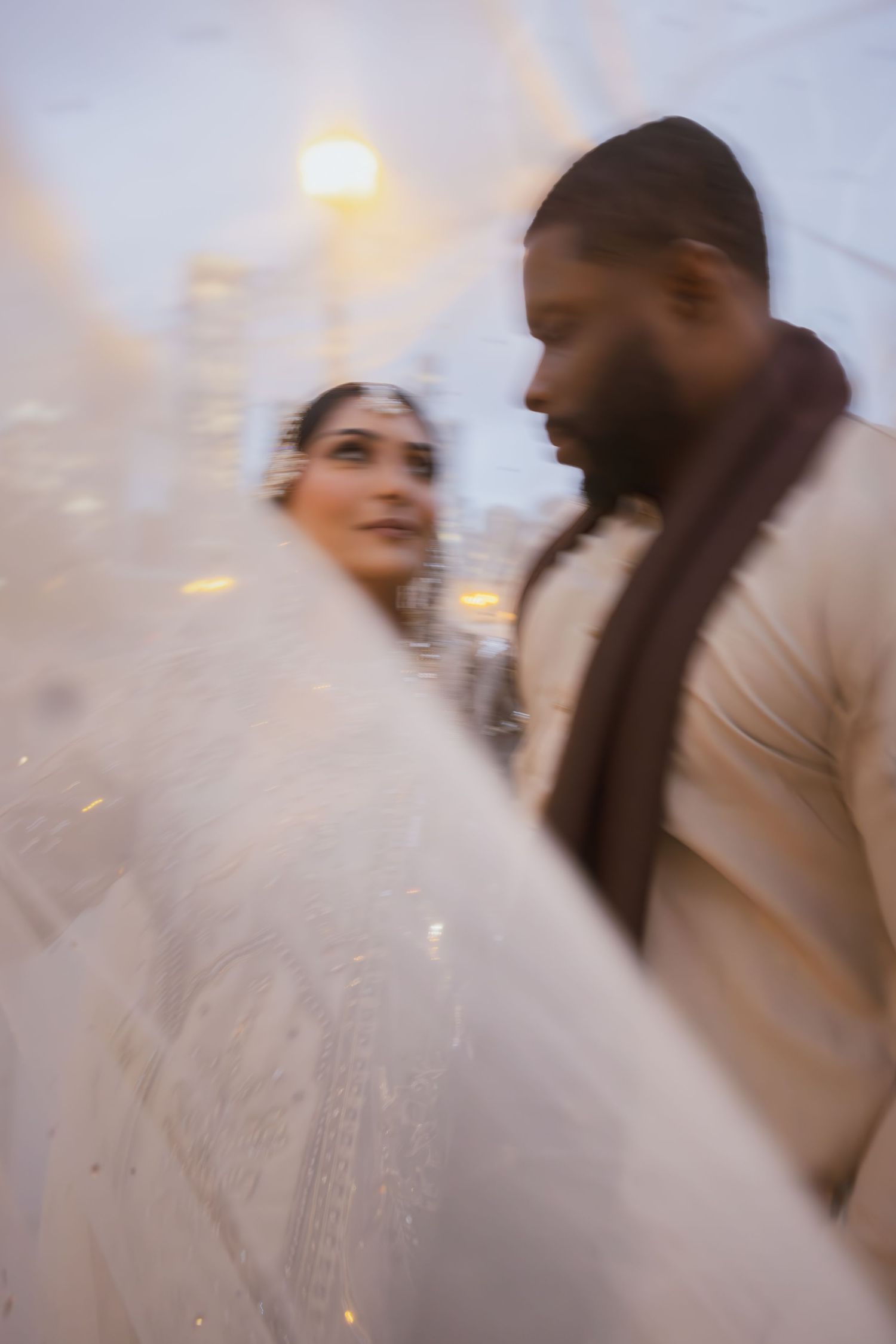 Blurred artistic shot of South Asian bride in white lehenga with groom in cream sherwani at wedding.