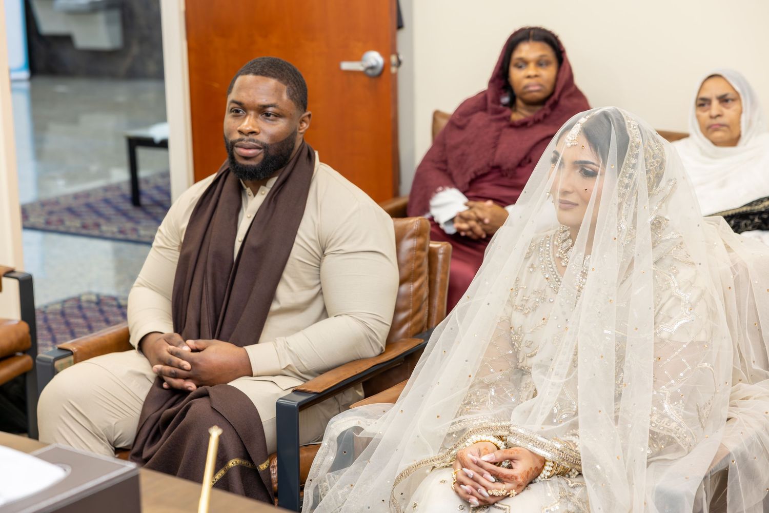 Interracial Muslim couple seated together during nikah ceremony with family members watching in background.