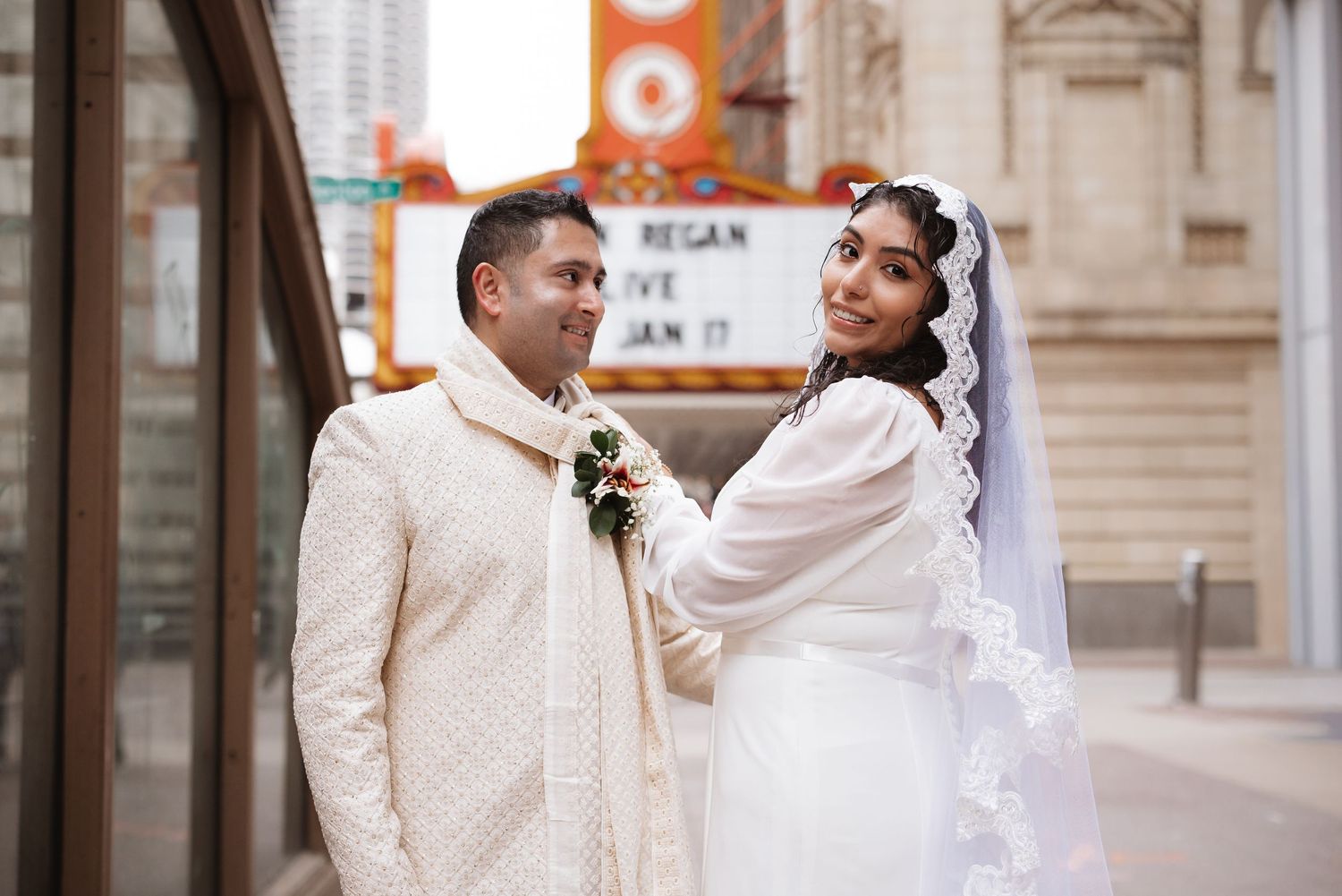 South Asian groom in embroidered sherwani and veiled bride share a tender moment outside theater.