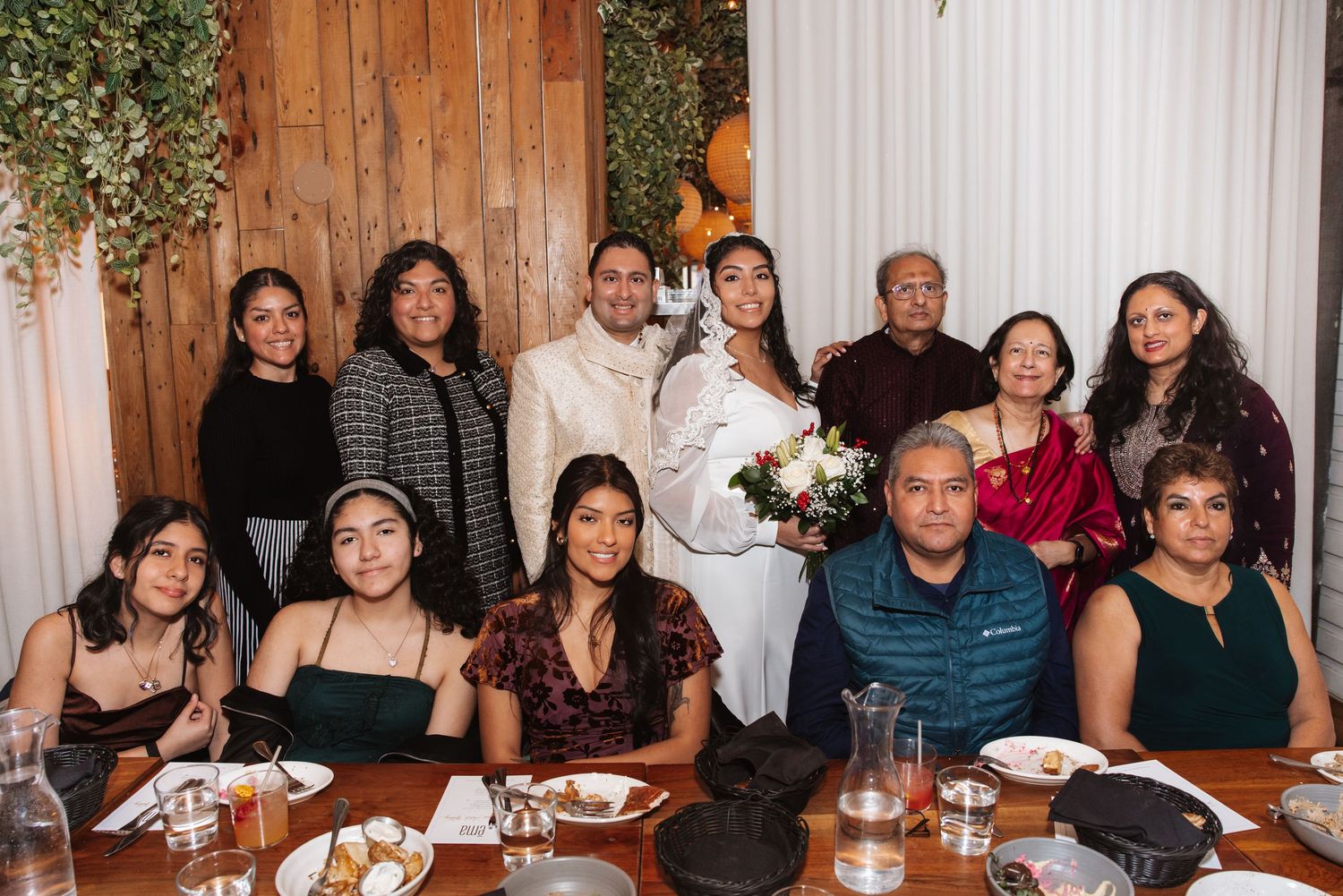 Wedding couple with bouquet posing with large family group at decorated reception dinner table in rustic restaurant setting.