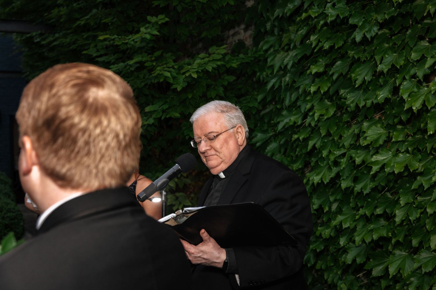 Officiant in black clergy attire reading from book at outdoor wedding ceremony with ivy wall background.