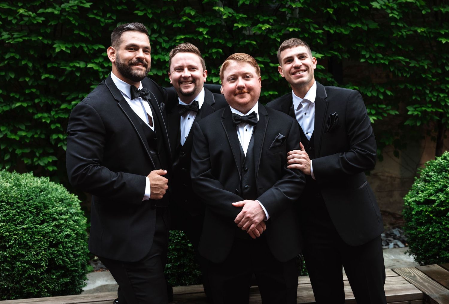 Four smiling groomsmen in black tuxedos posing together outdoors near manicured hedges and an ivy wall.