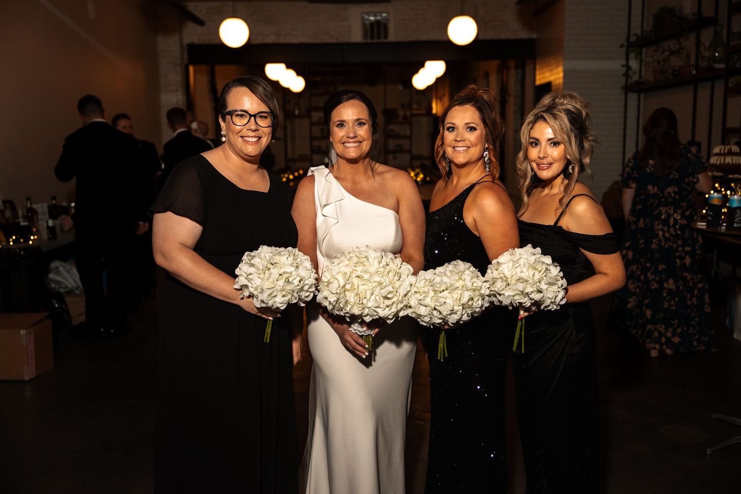 Bride in white gown poses with three bridesmaids in black dresses holding white hydrangea bouquets.