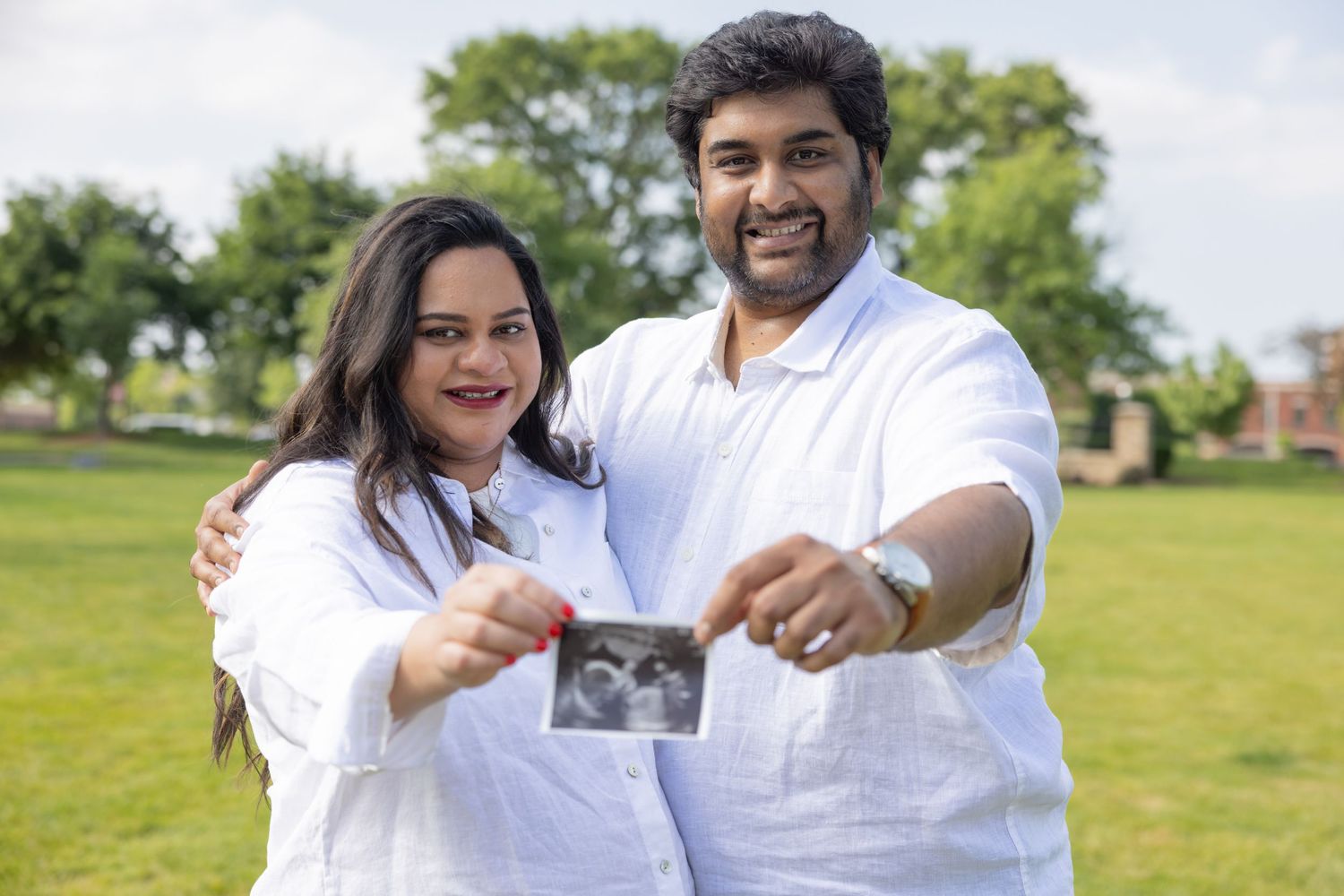 Smiling Indian couple in white outfits holding ultrasound photo in a green park.