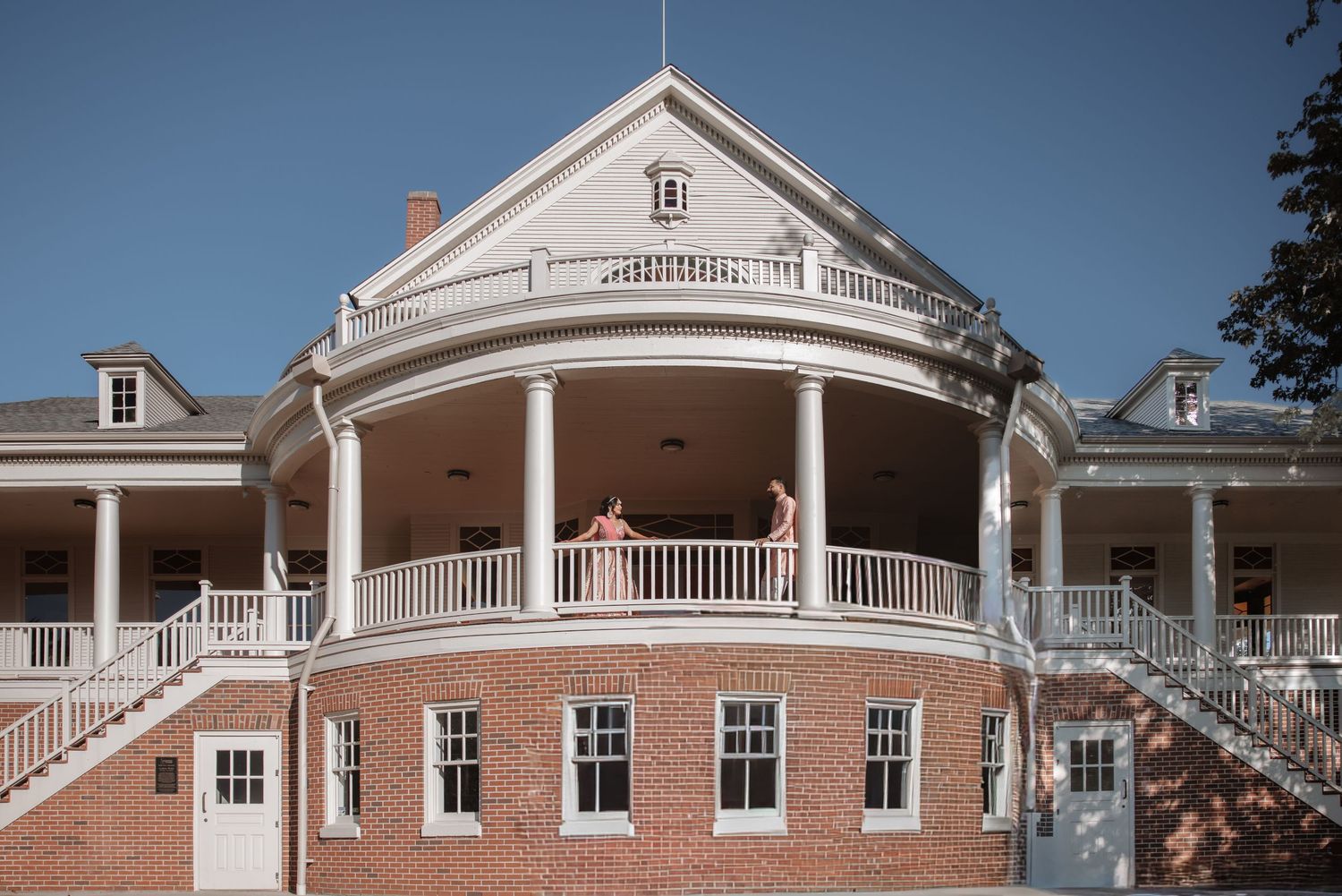 Elegant brick colonial building with white wraparound balcony and columns photographed on a clear sunny day.