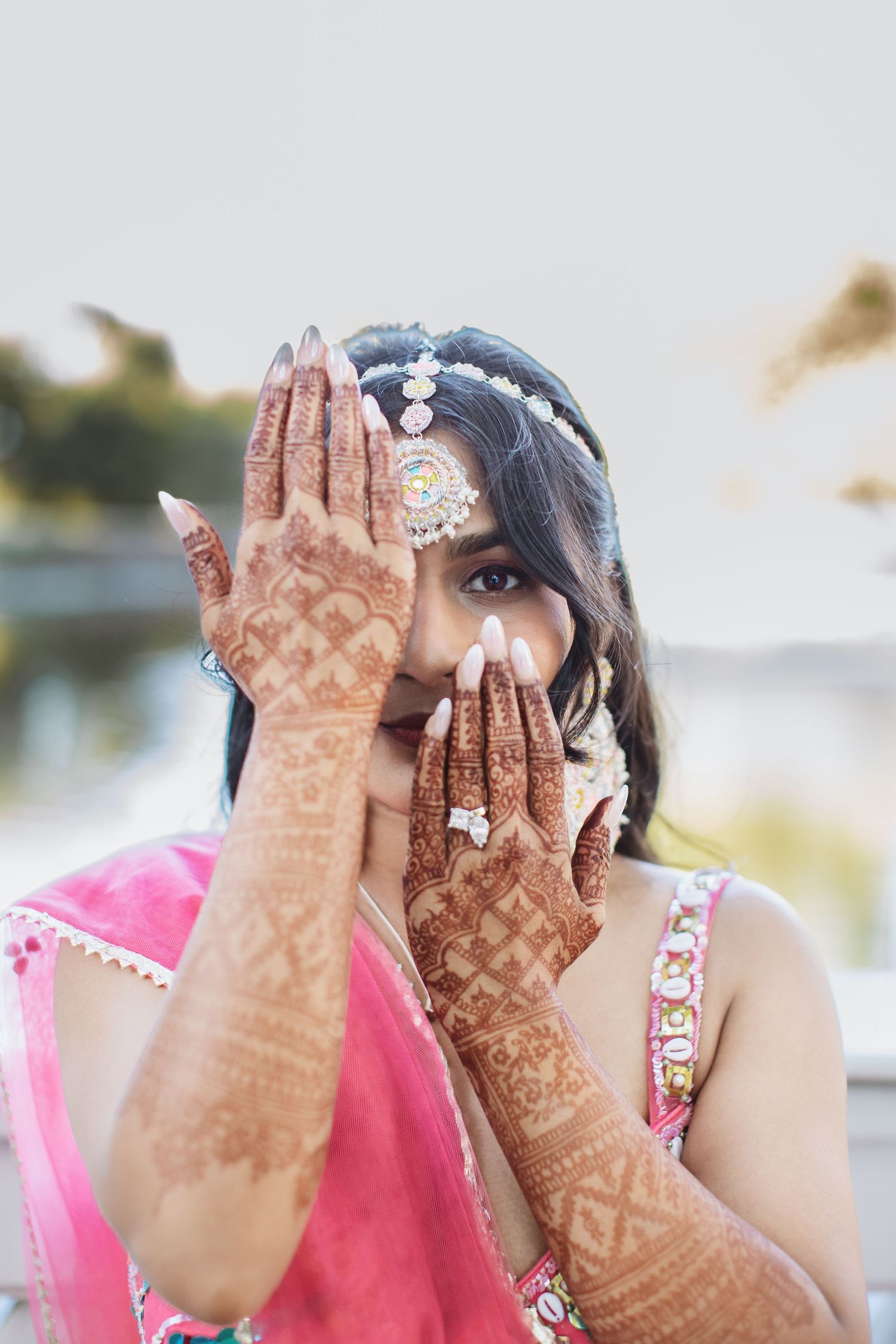 Indian bride in pink lehenga showcases intricate mehndi henna designs on both hands with bridal jewelry.