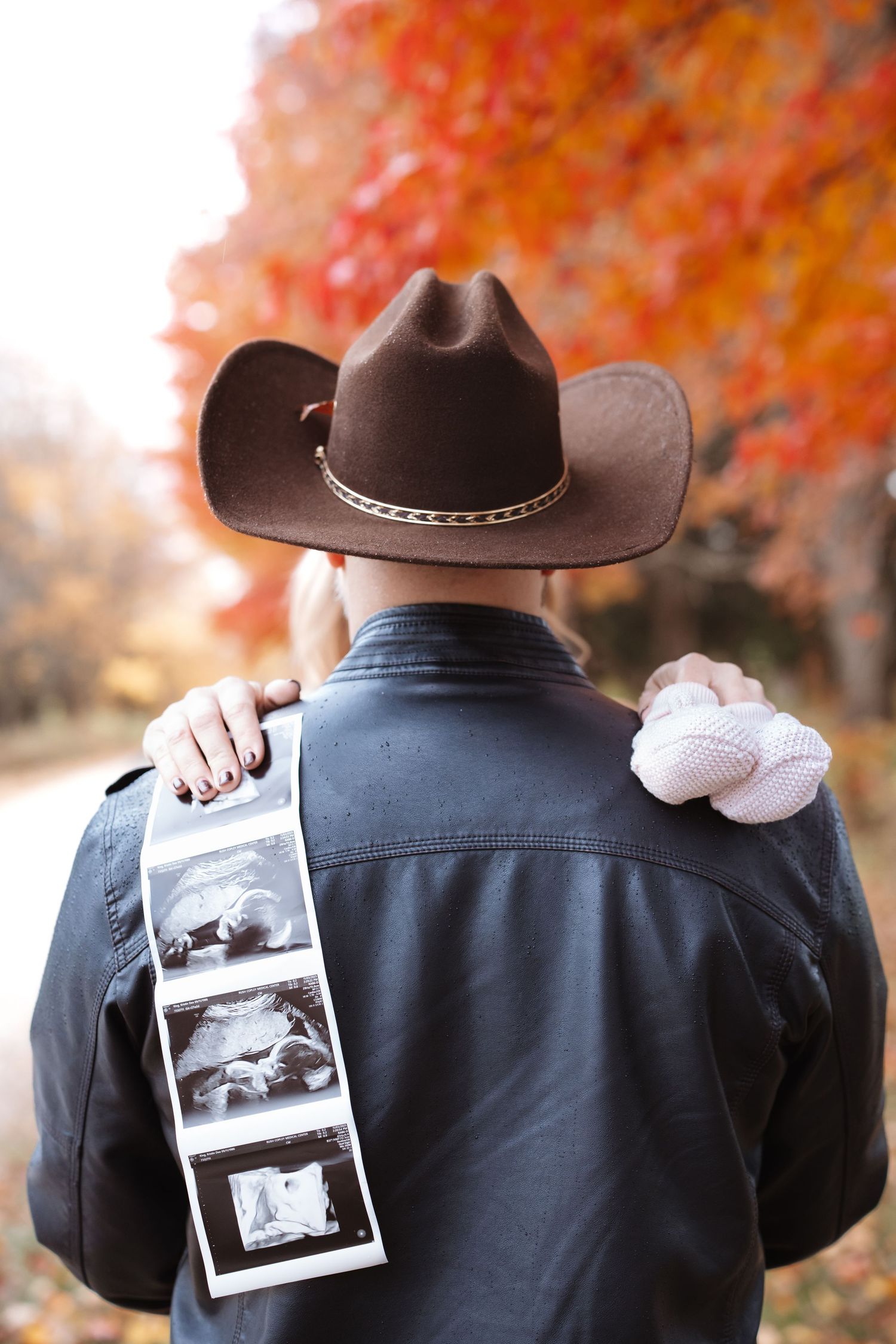 Rear view of man in brown cowboy hat holding ultrasound strip with tiny baby shoes in fall setting.