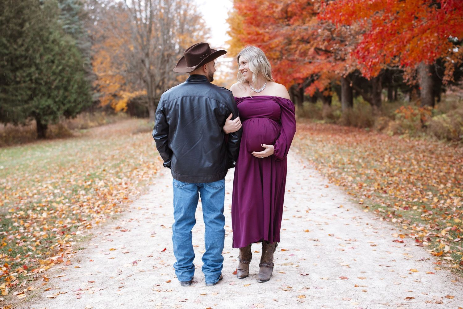 Couple walking on autumn path, pregnant woman in purple dress with partner in cowboy hat and leather jacket.