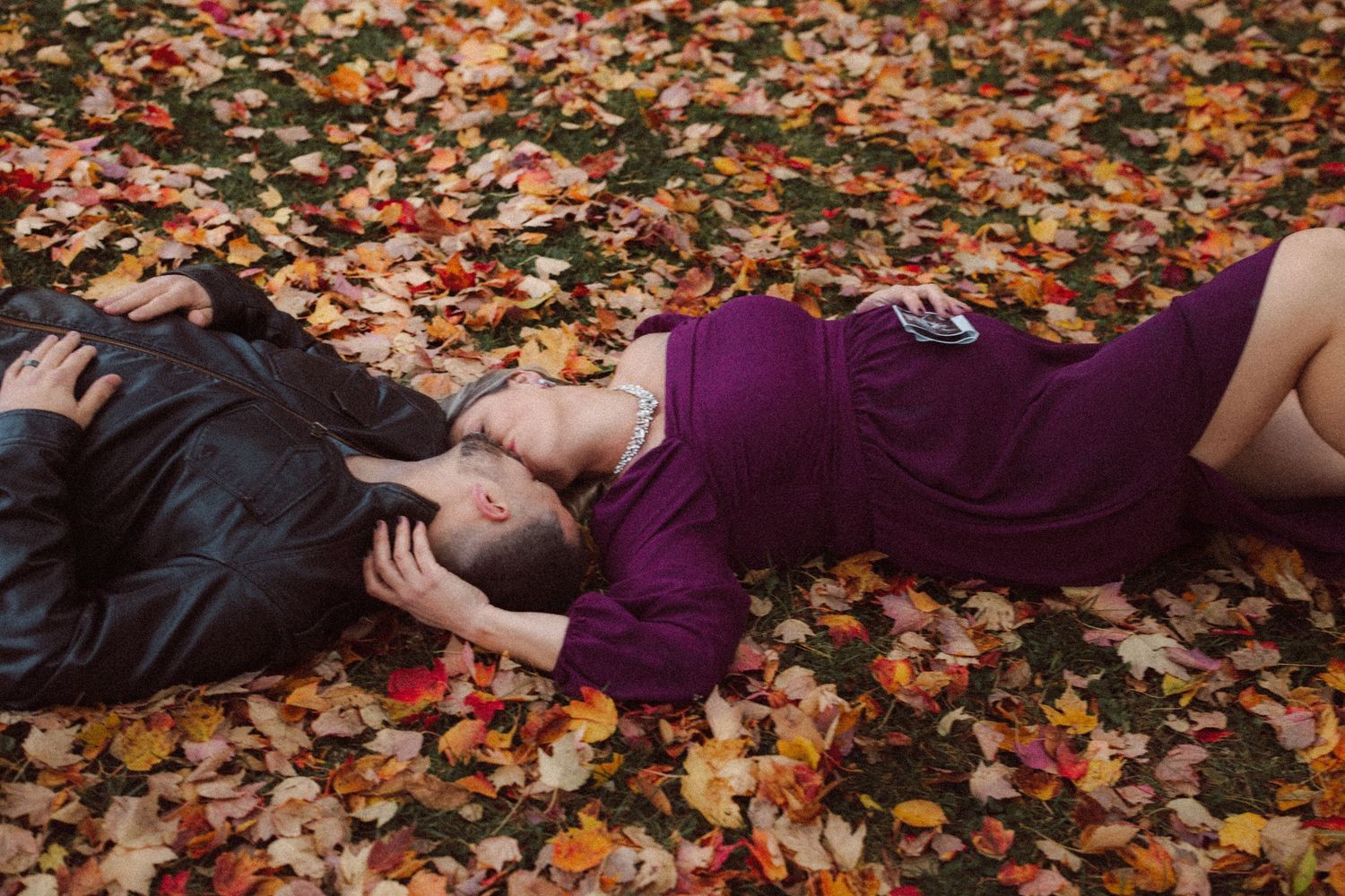 Couple lying in colorful fall leaves, pregnant woman in purple dress resting close to her partner.