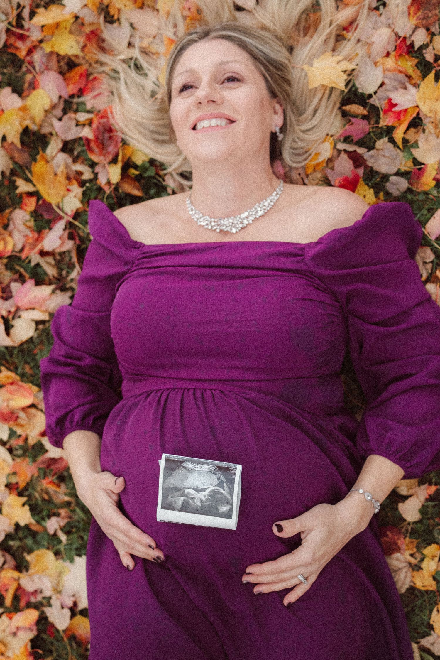 Smiling pregnant woman in purple dress lying in autumn leaves holding ultrasound photo on her belly.
