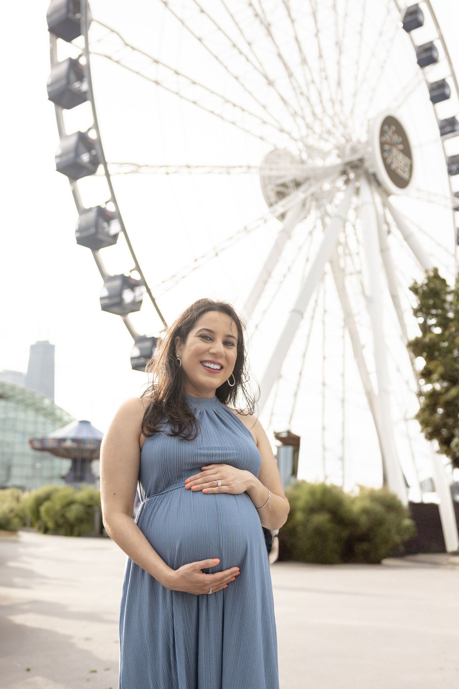 Smiling pregnant woman in blue dress poses by a Ferris wheel.