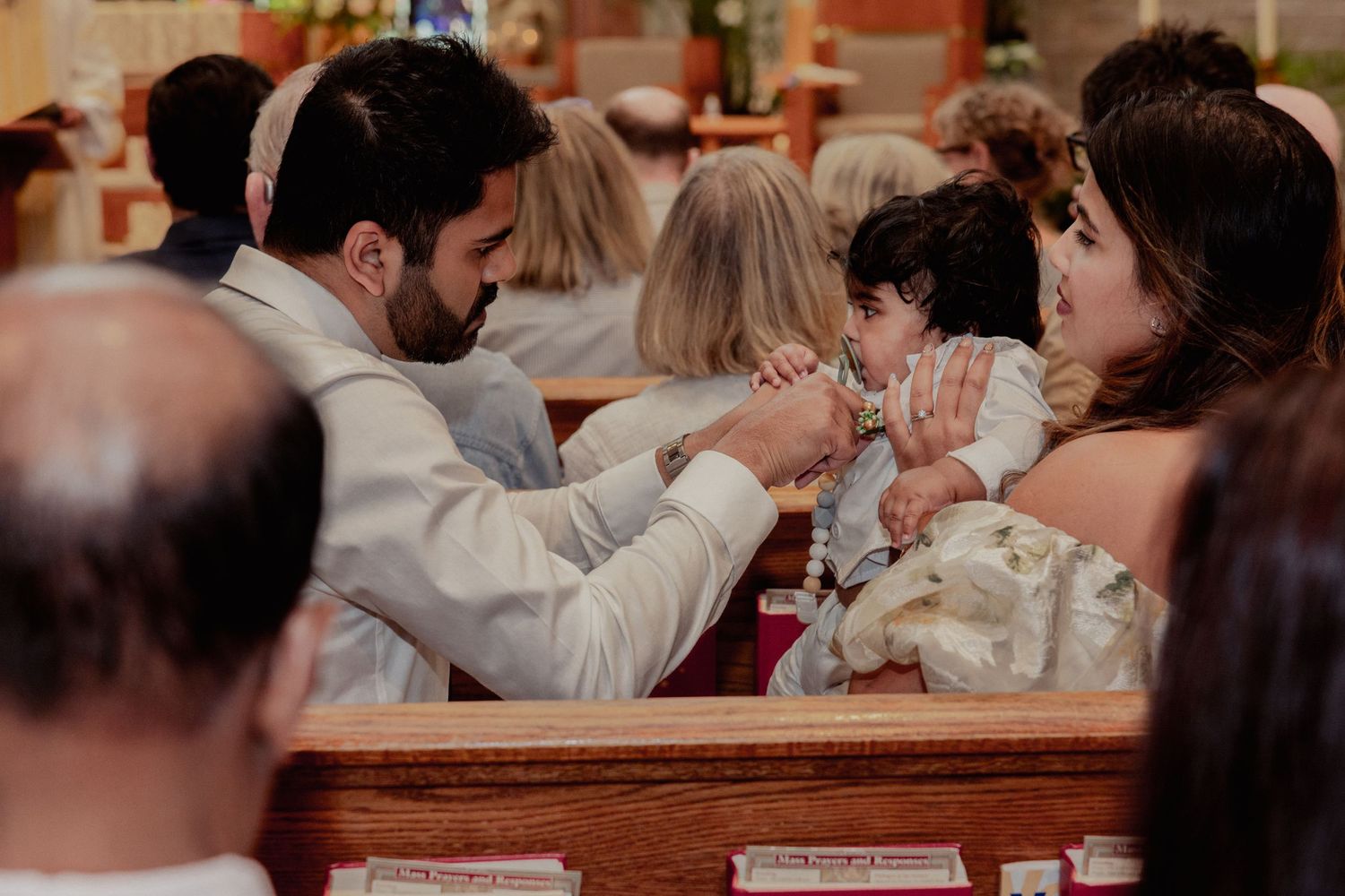 Parents interact lovingly with baby during Catholic baptism ceremony in church pew.