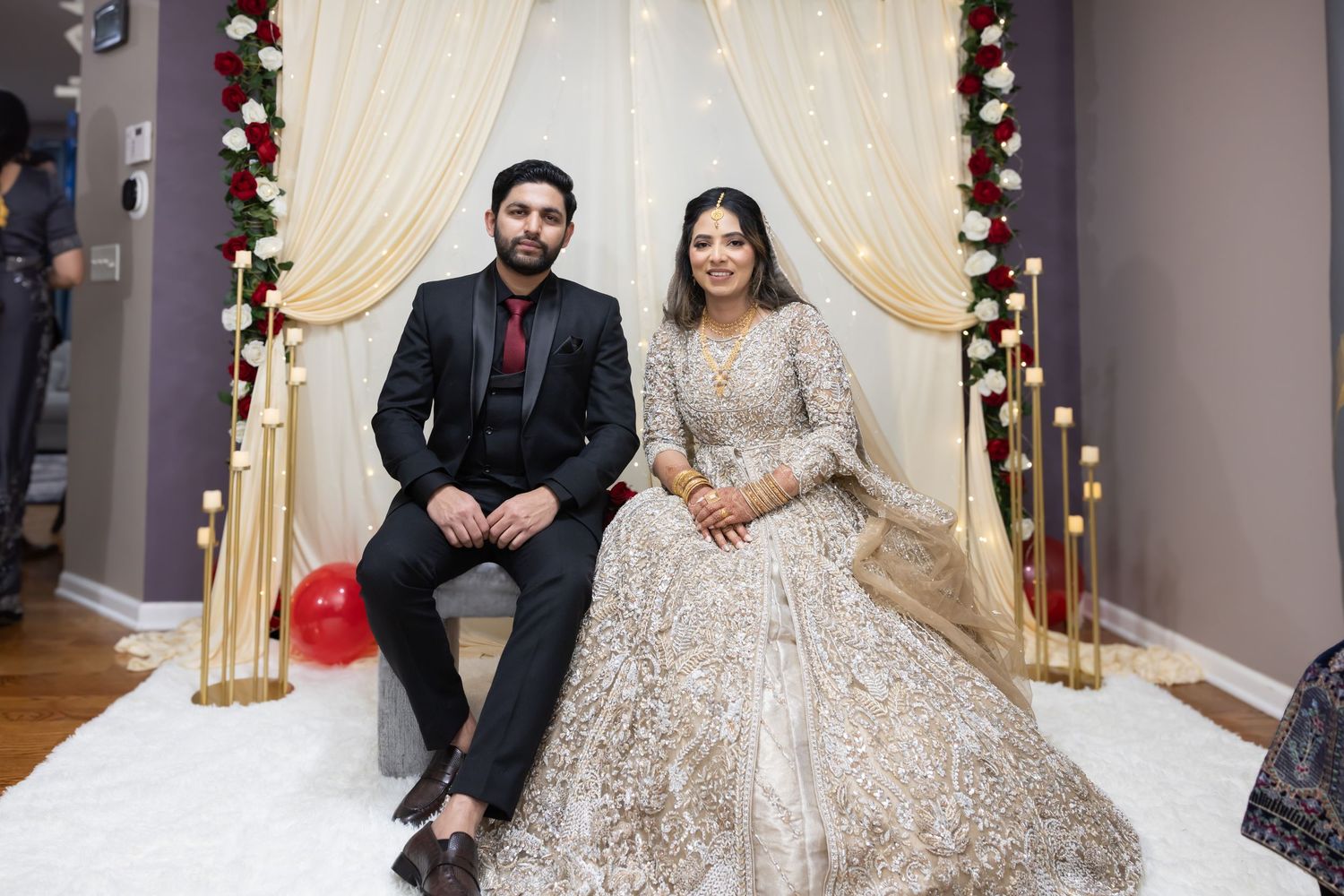 South Asian bride and groom seated at decorated wedding mandap with floral backdrop.