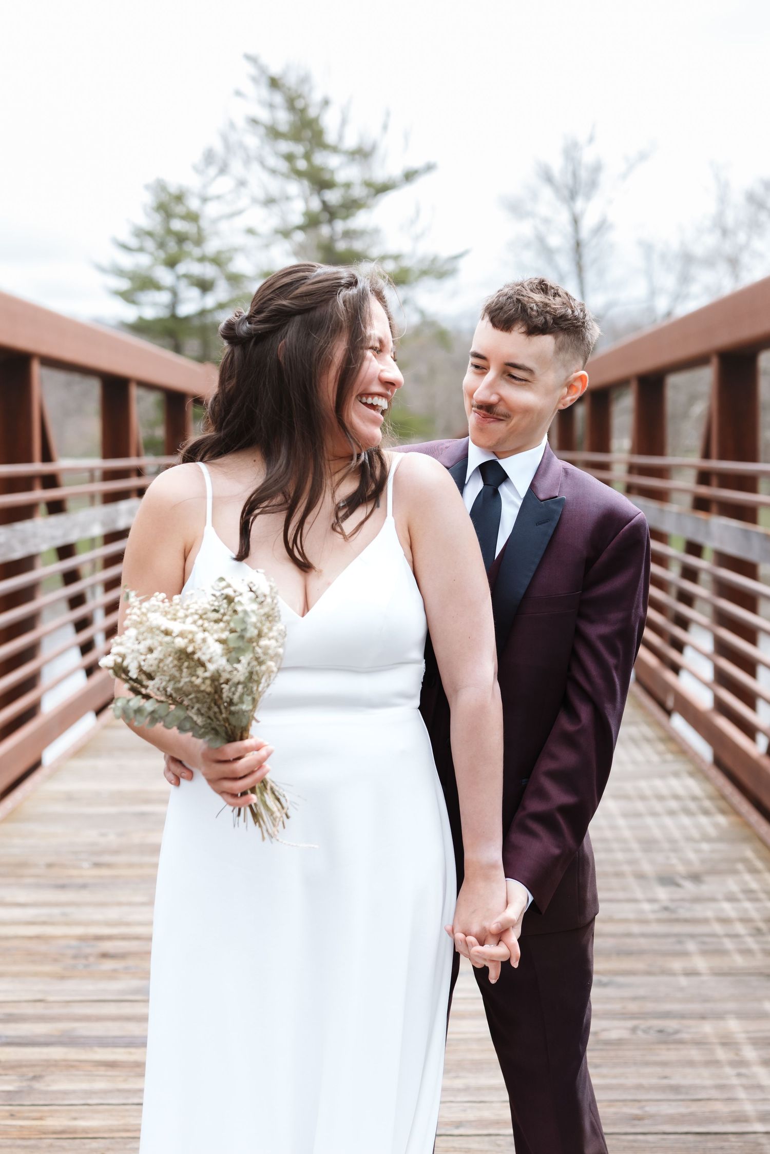 Couple holding hands on a wooden bridge, bride in white gown with bouquet, groom in burgundy suit, smiling at each other.