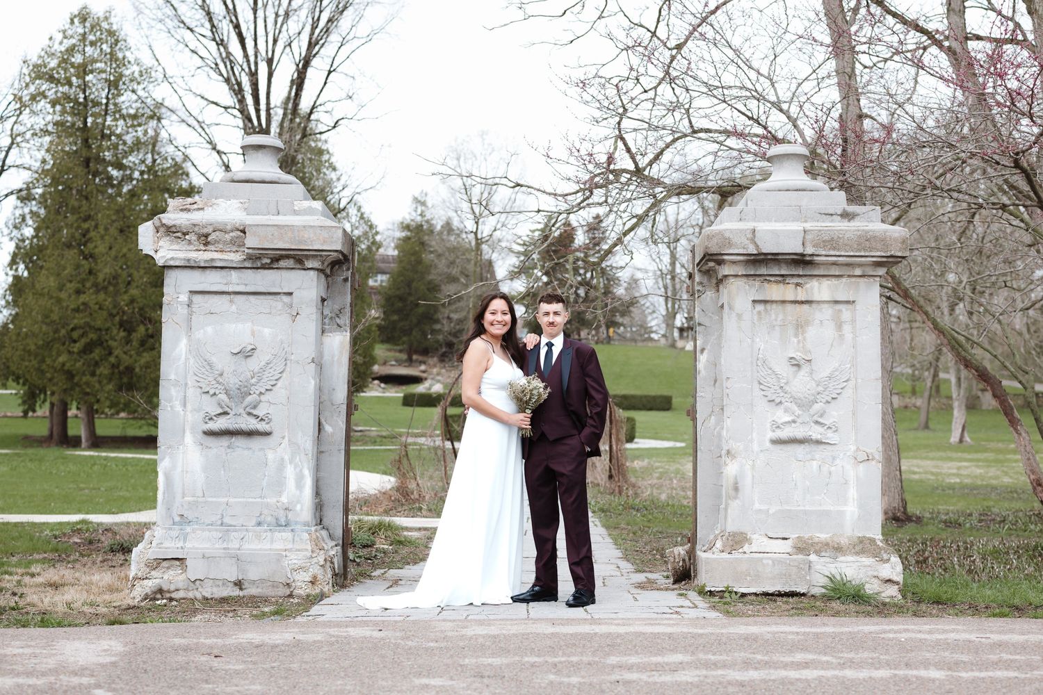 Bride and groom posing between ornate stone gate pillars in a lush green park on their wedding day.