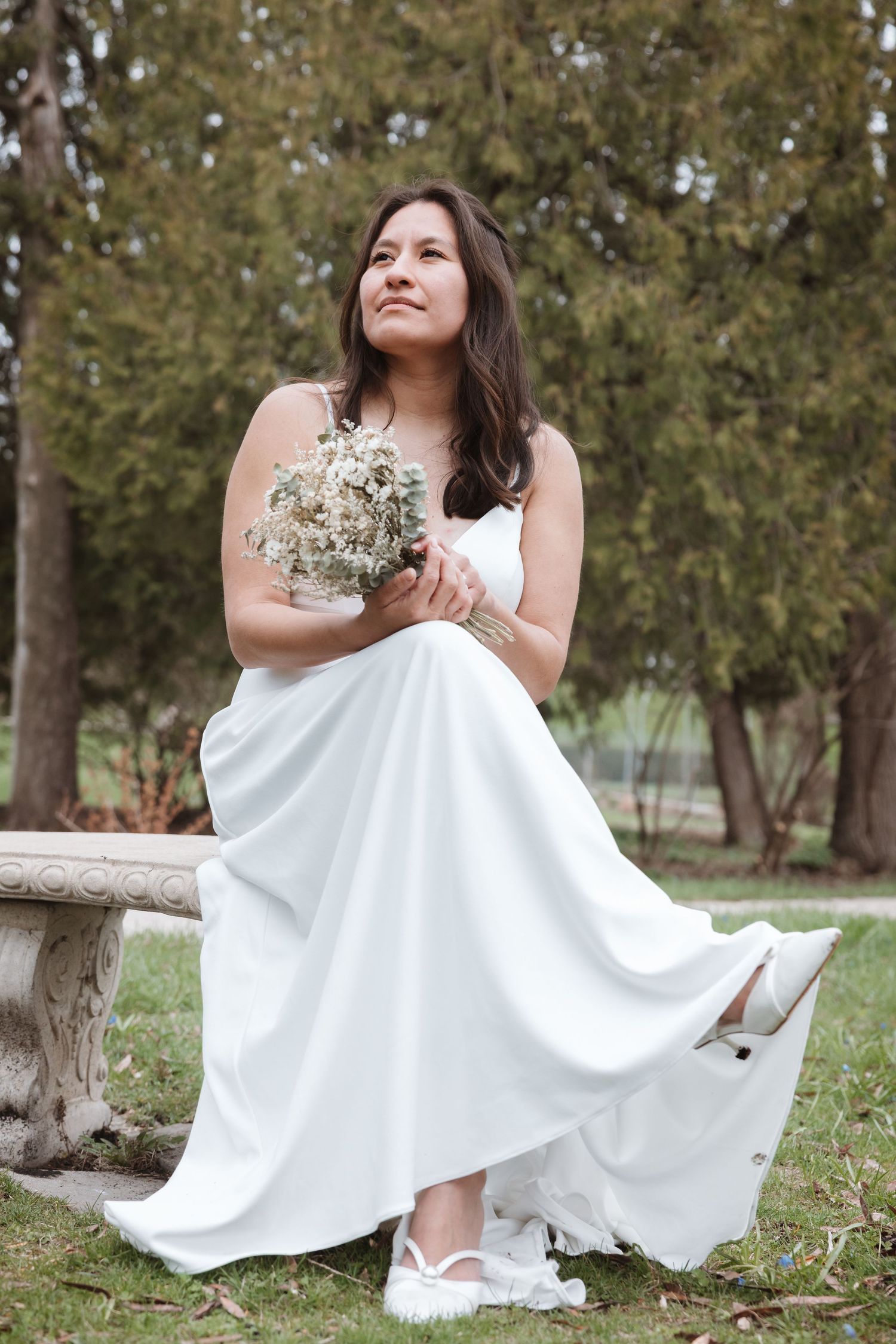 Bride in white gown holding dried flower bouquet, seated on stone bench in lush green park, looking up dreamily.