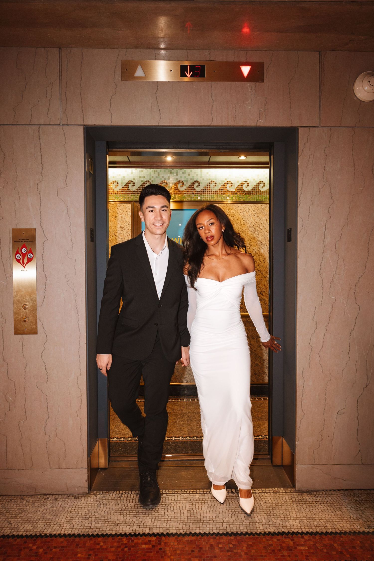Elegant couple stepping out of ornate gold elevator, man in black suit and woman in white fitted dress.Chicago Wedding Photographer