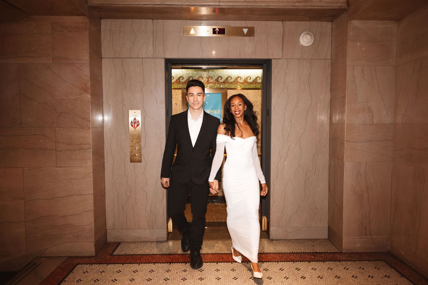 Elegant couple holding hands walking out of ornate elevator, man in black suit, woman in white gown.Chicago Wedding Photographer