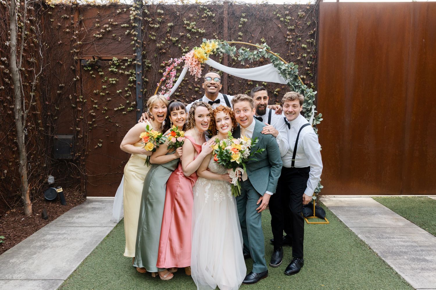 Bride and groom celebrate joyfully with wedding party in front of floral ceremony arch.