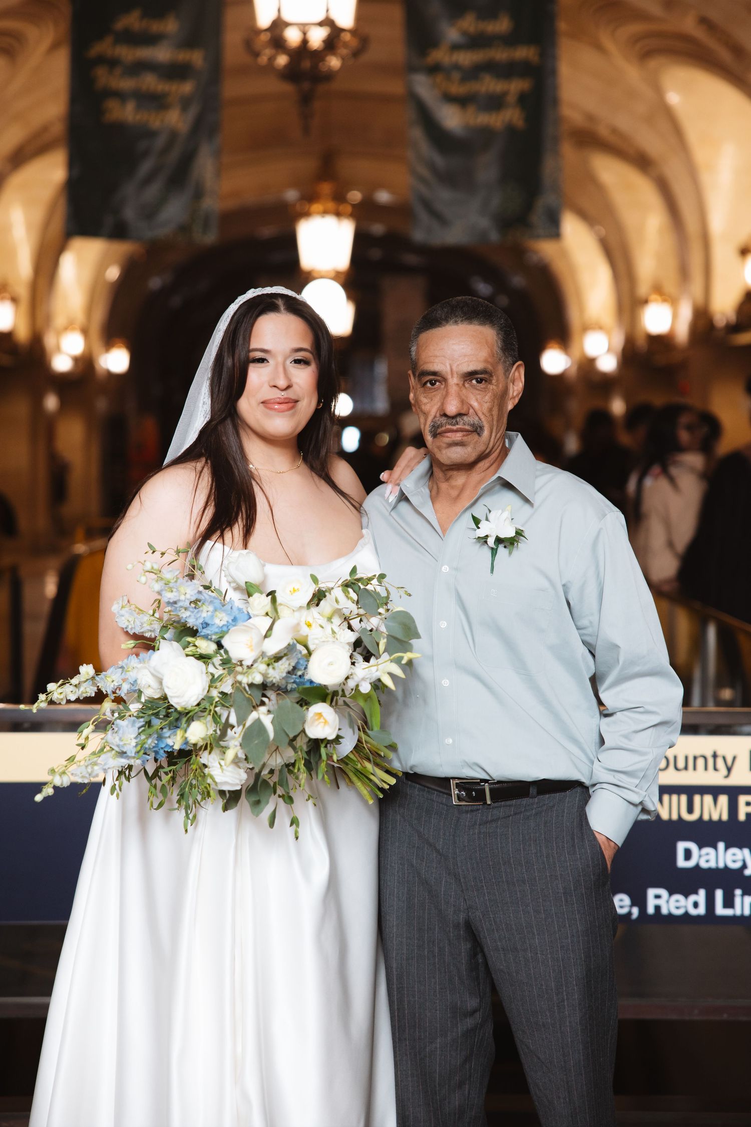 Bride with veil and bouquet poses with older man wearing boutonniere in grand hall.