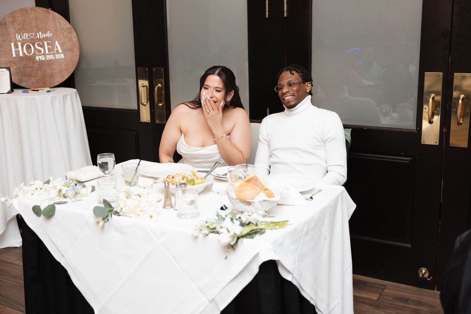 Newlyweds laugh together at sweetheart table decorated with white flowers at reception.