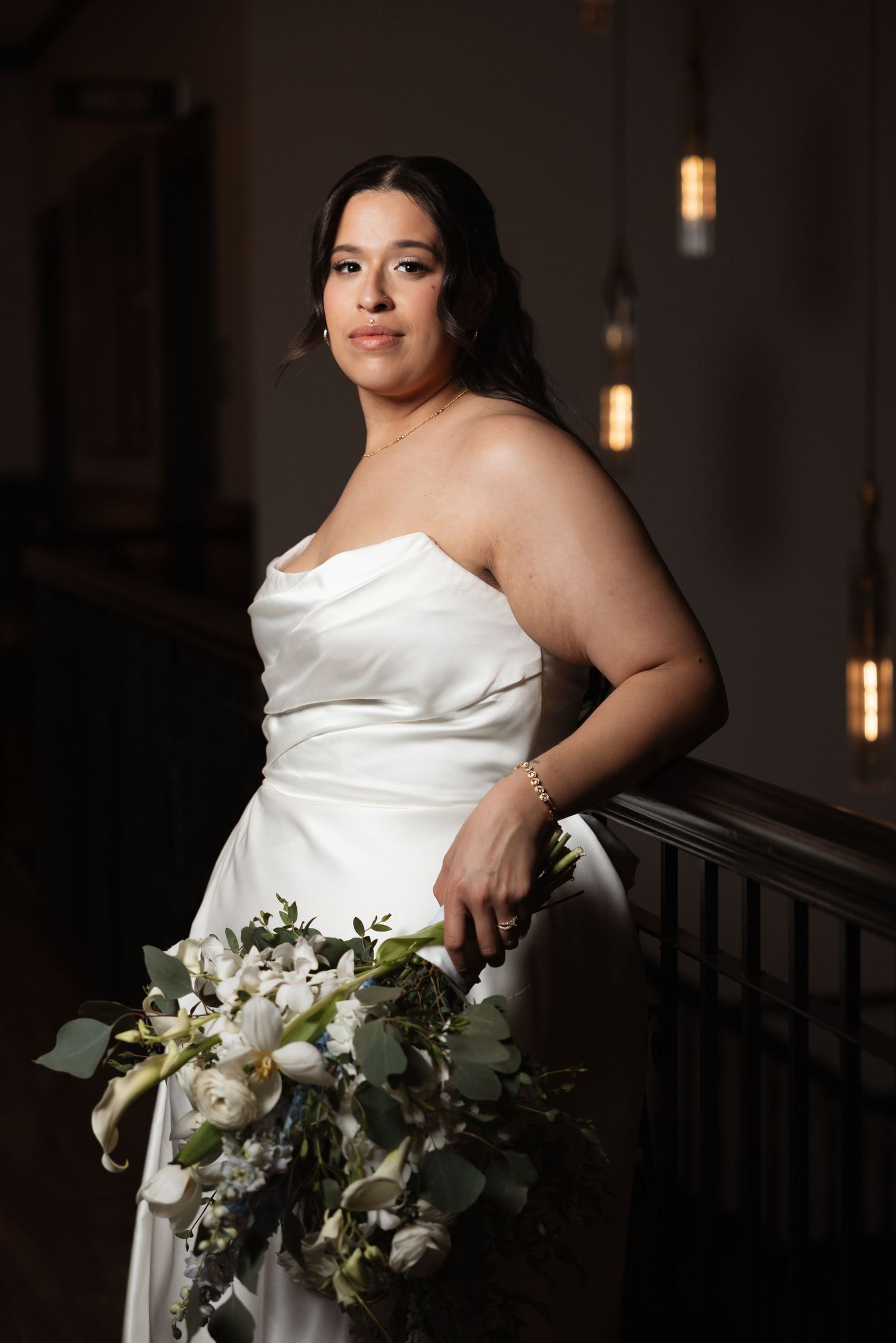 Bride in strapless white satin gown holding white floral bouquet posing in elegant venue.
