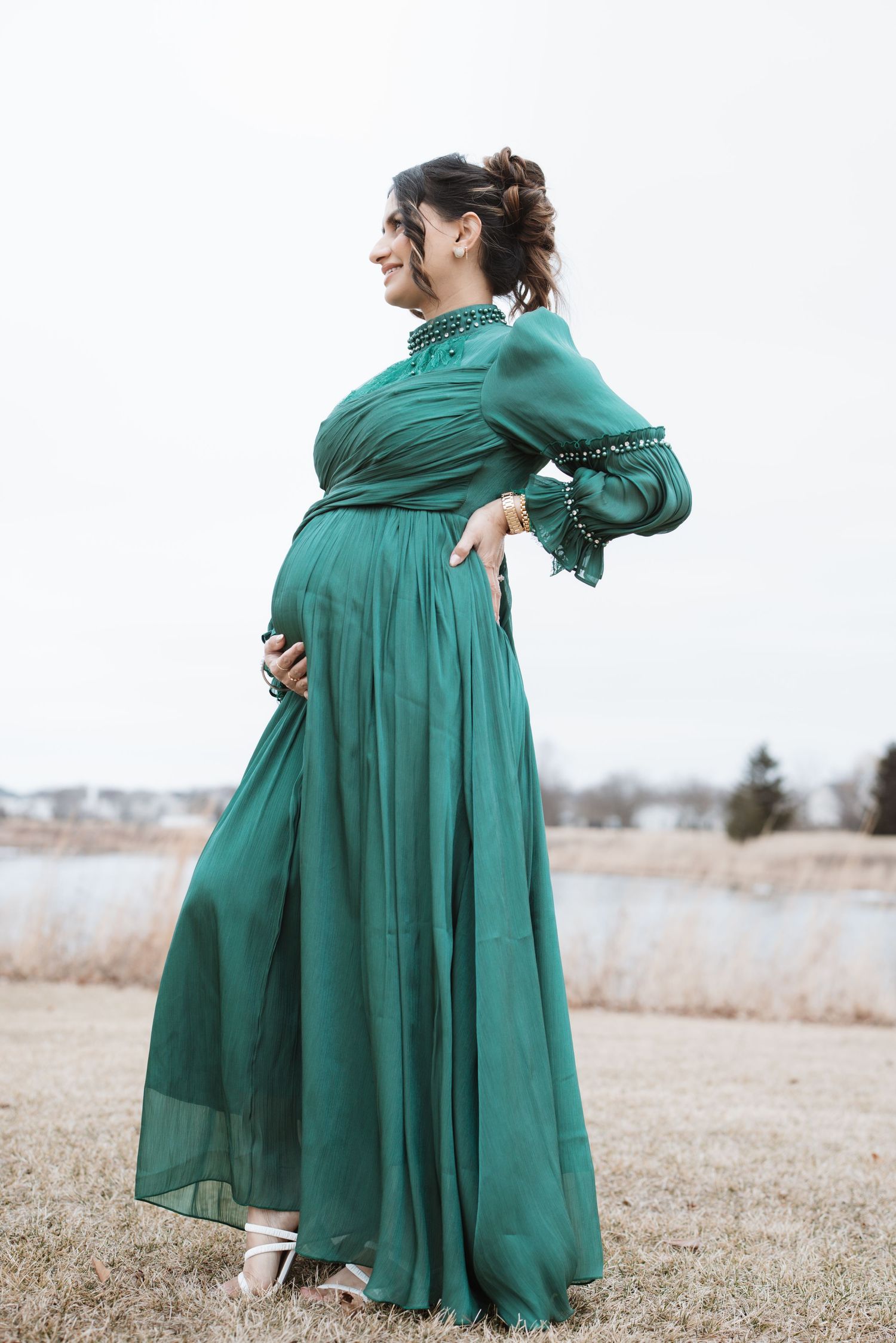 Pregnant woman in elegant green maxi dress holding baby bump outdoors near lake.