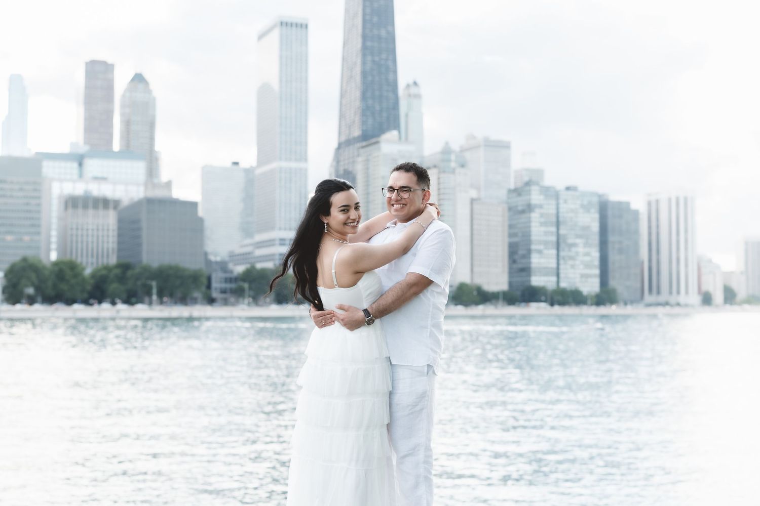 Couple embracing in white outfits by Chicago waterfront with city skyline backdrop on a cloudy day.